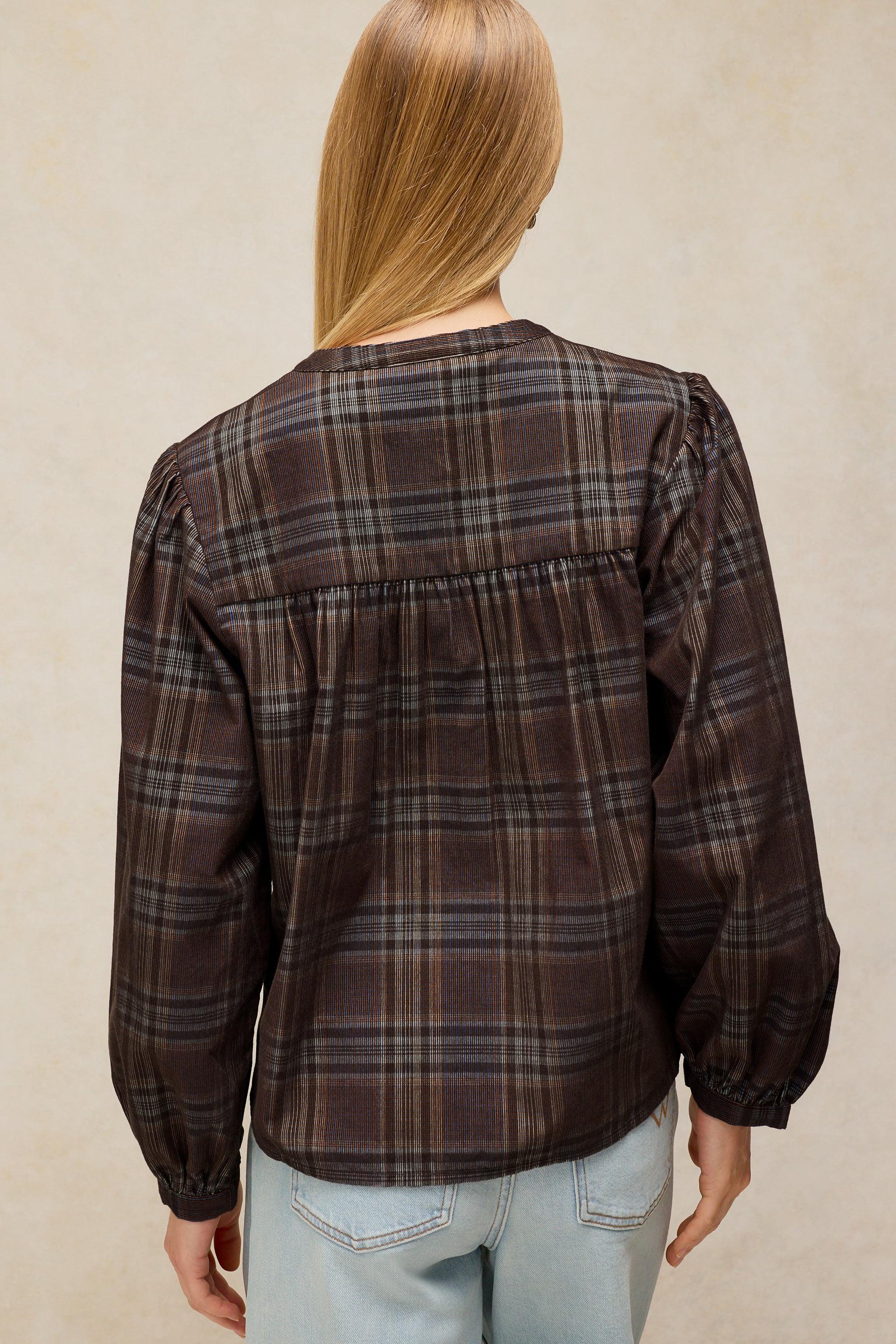 A woman viewed from the back wearing a dark brown and black plaid blouse with long balloon sleeves and a gathered waist seam, paired with light blue jeans against a textured beige background.