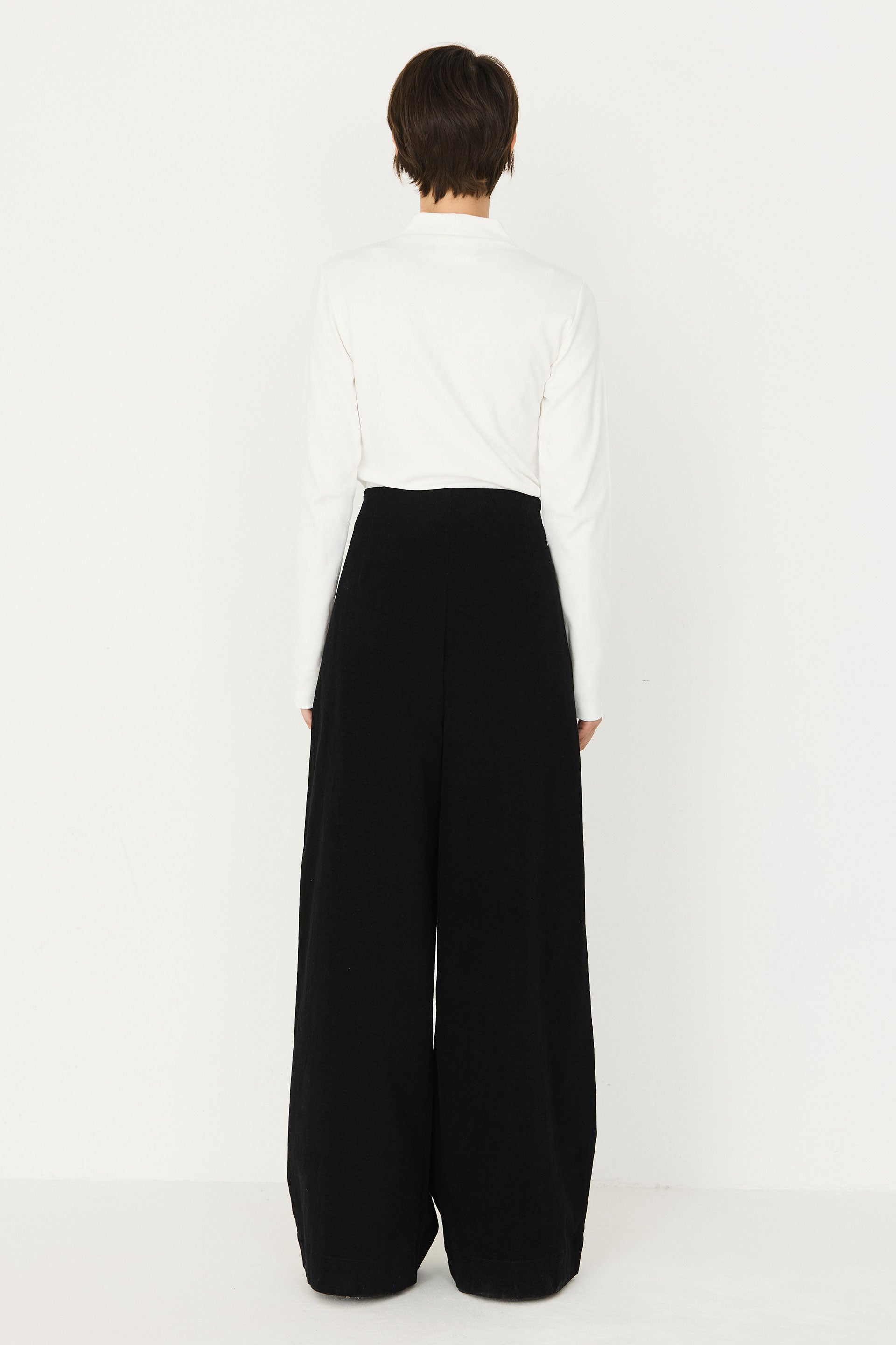 Back view of a woman wearing a white long-sleeve organic cotton top by SKFK and black wide-leg trousers.