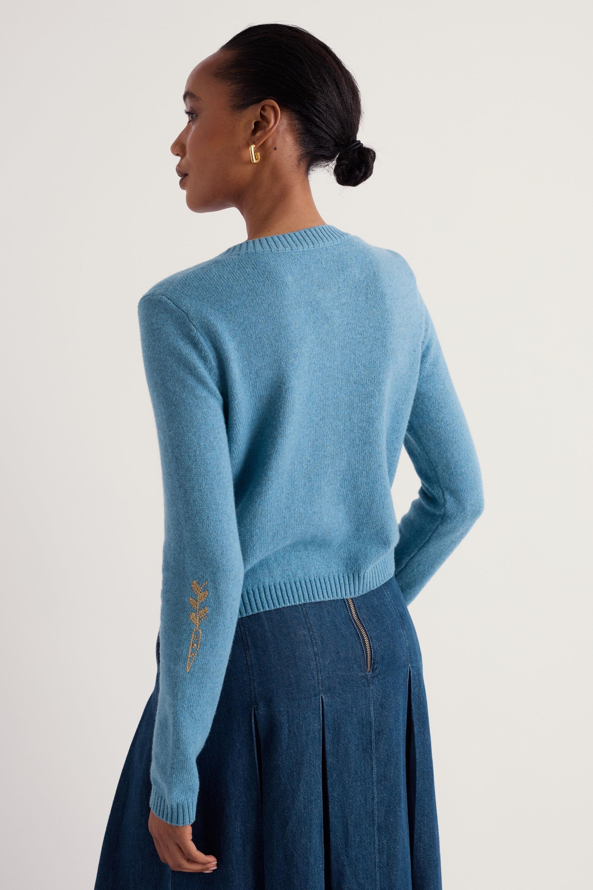 A woman shown from the back wearing a light blue knit sweater with gold embroidered details on the sleeve and a dark blue pleated skirt, against a plain light gray studio background.
