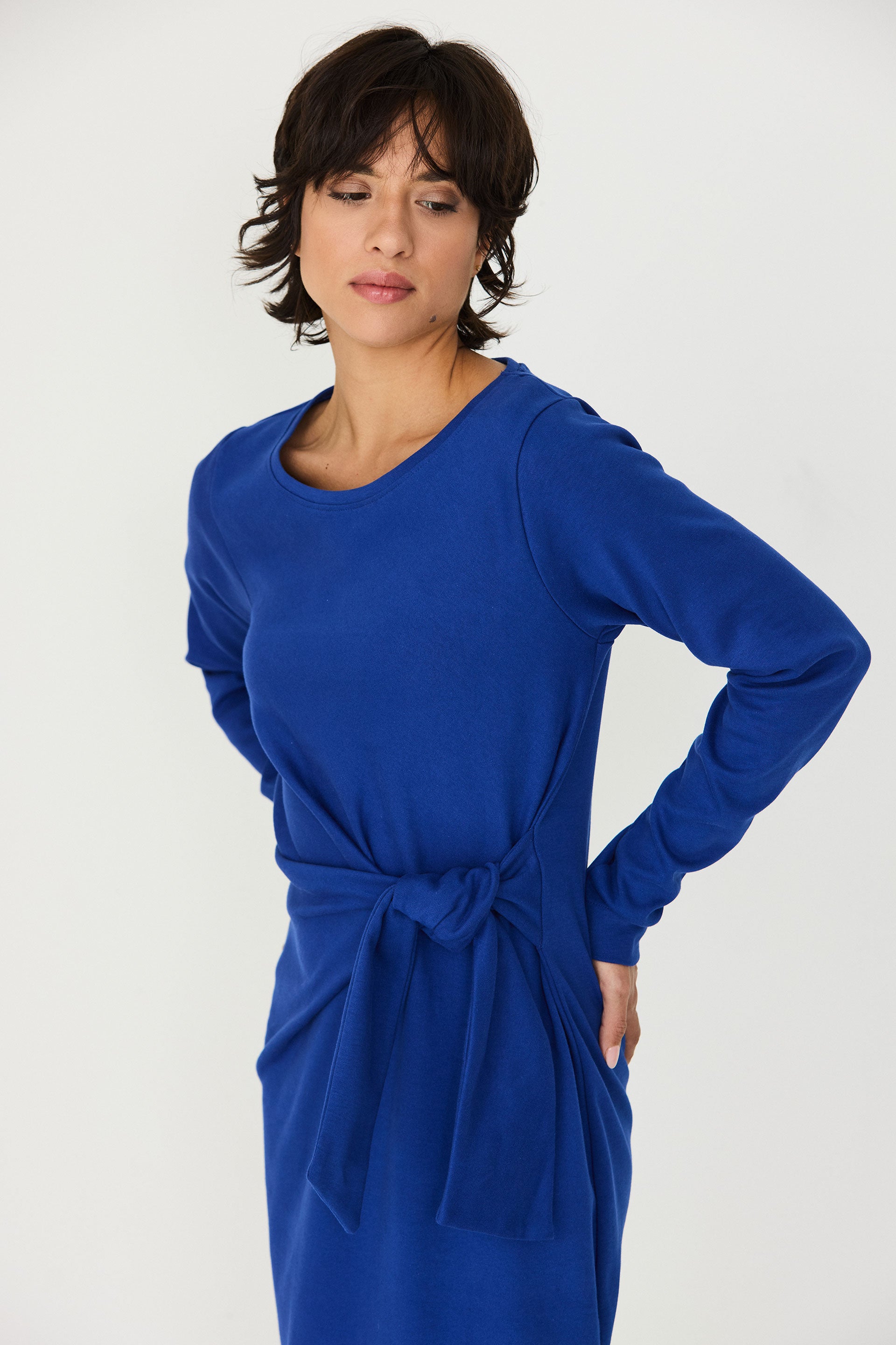 A woman modeling a long-sleeve blue dress with a tied waist belt, shown against a plain light gray studio background.