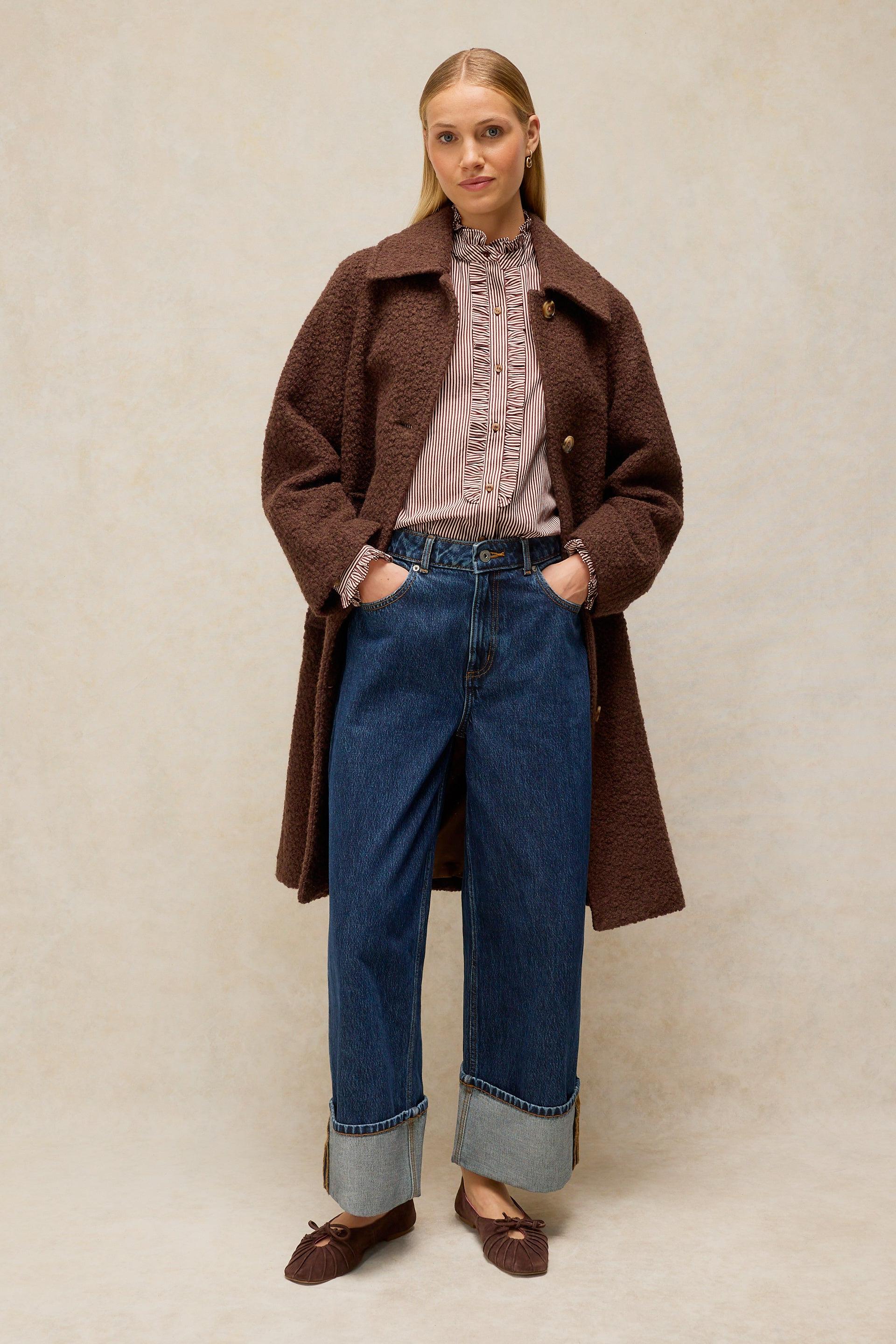 A woman stands against a neutral beige studio background wearing a dark brown textured coat over a dusty pink ruffled blouse, blue wide-leg jeans with cuffed hems, and brown loafers.