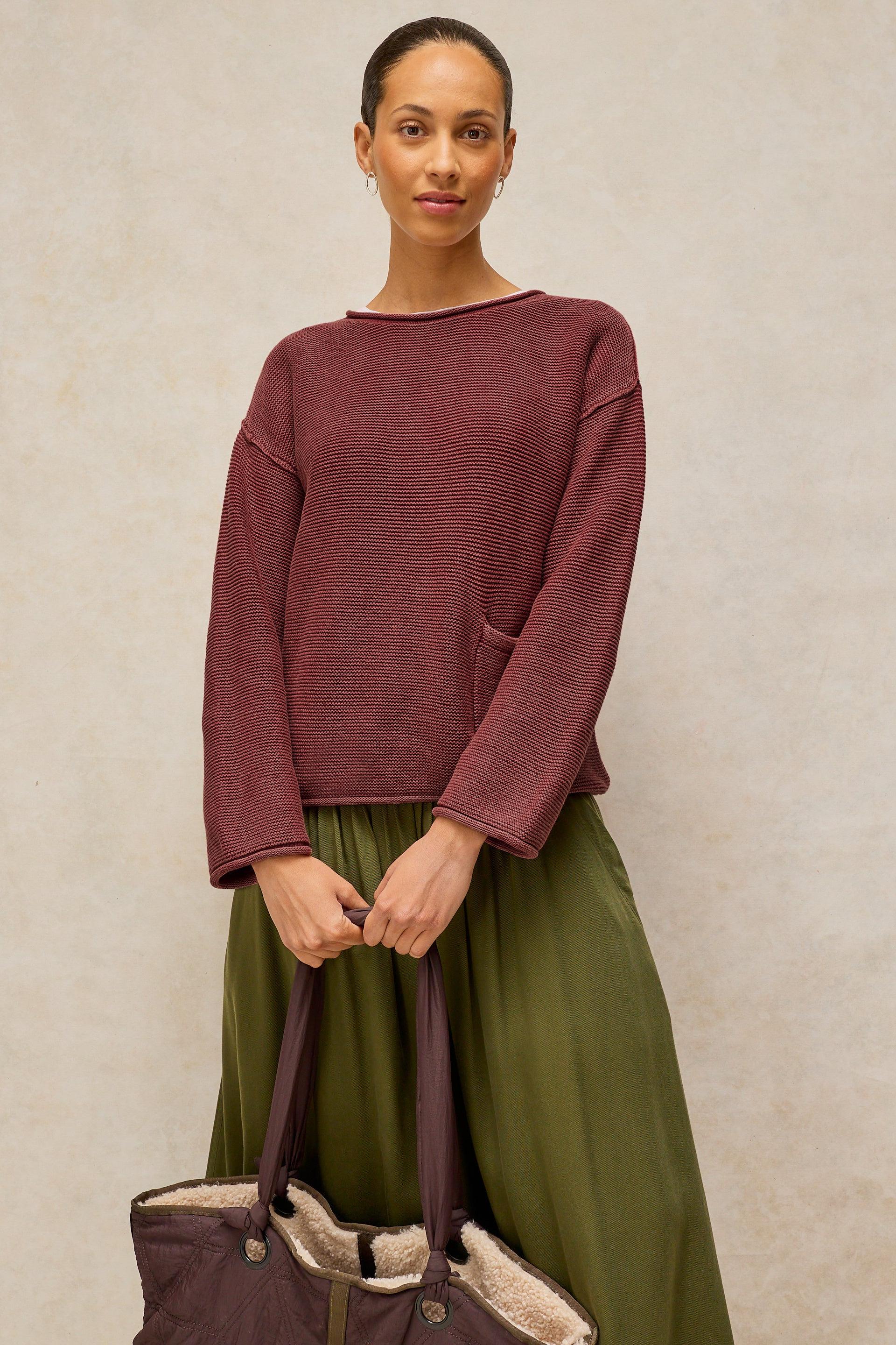 A woman stands against a textured beige wall wearing a ribbed long-sleeve boat-neck sweater in a muted burgundy shade, paired with an olive green skirt, and holding a dark plum tote bag with a light sherpa lining.