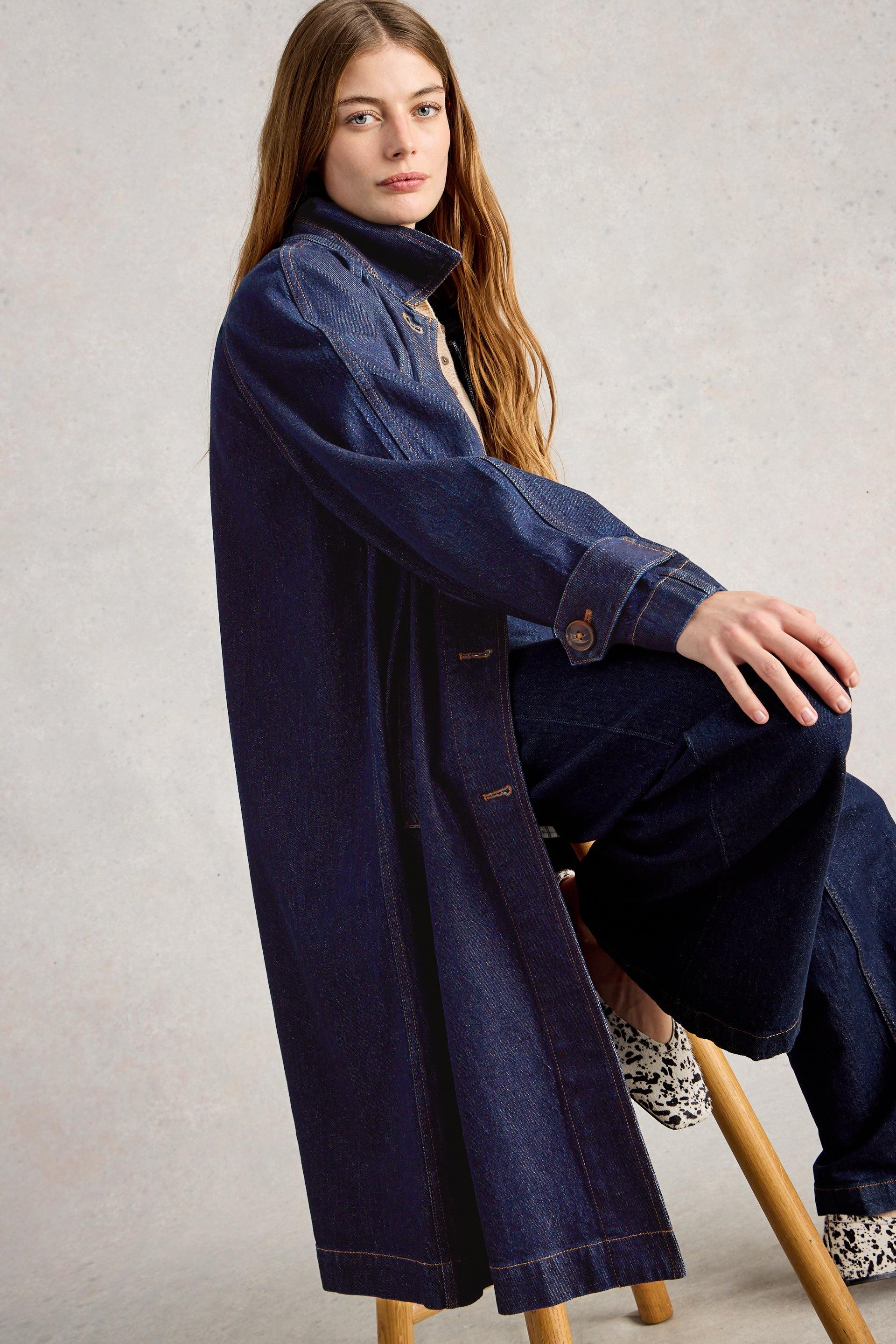 A woman wearing a long dark blue denim coat and organic cotton clothing sitting on a wooden stool