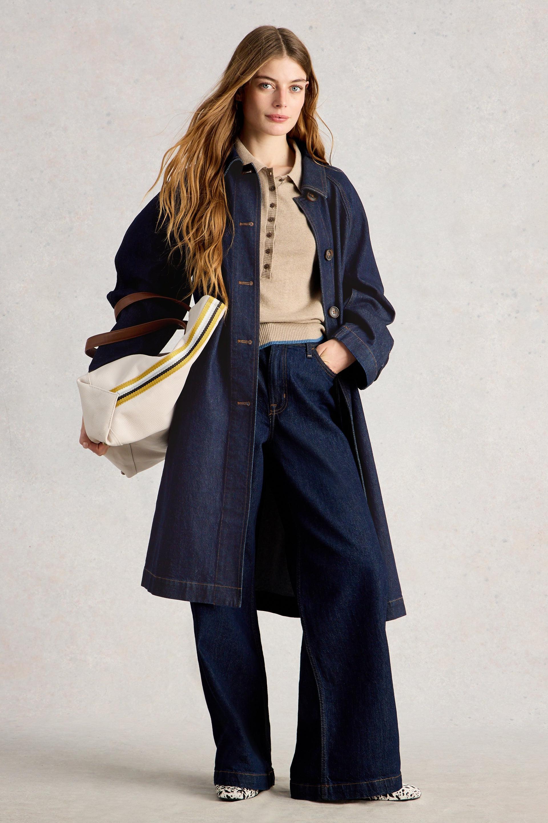 Woman wearing a long navy coat holding a white bag with a brown handle on a light background