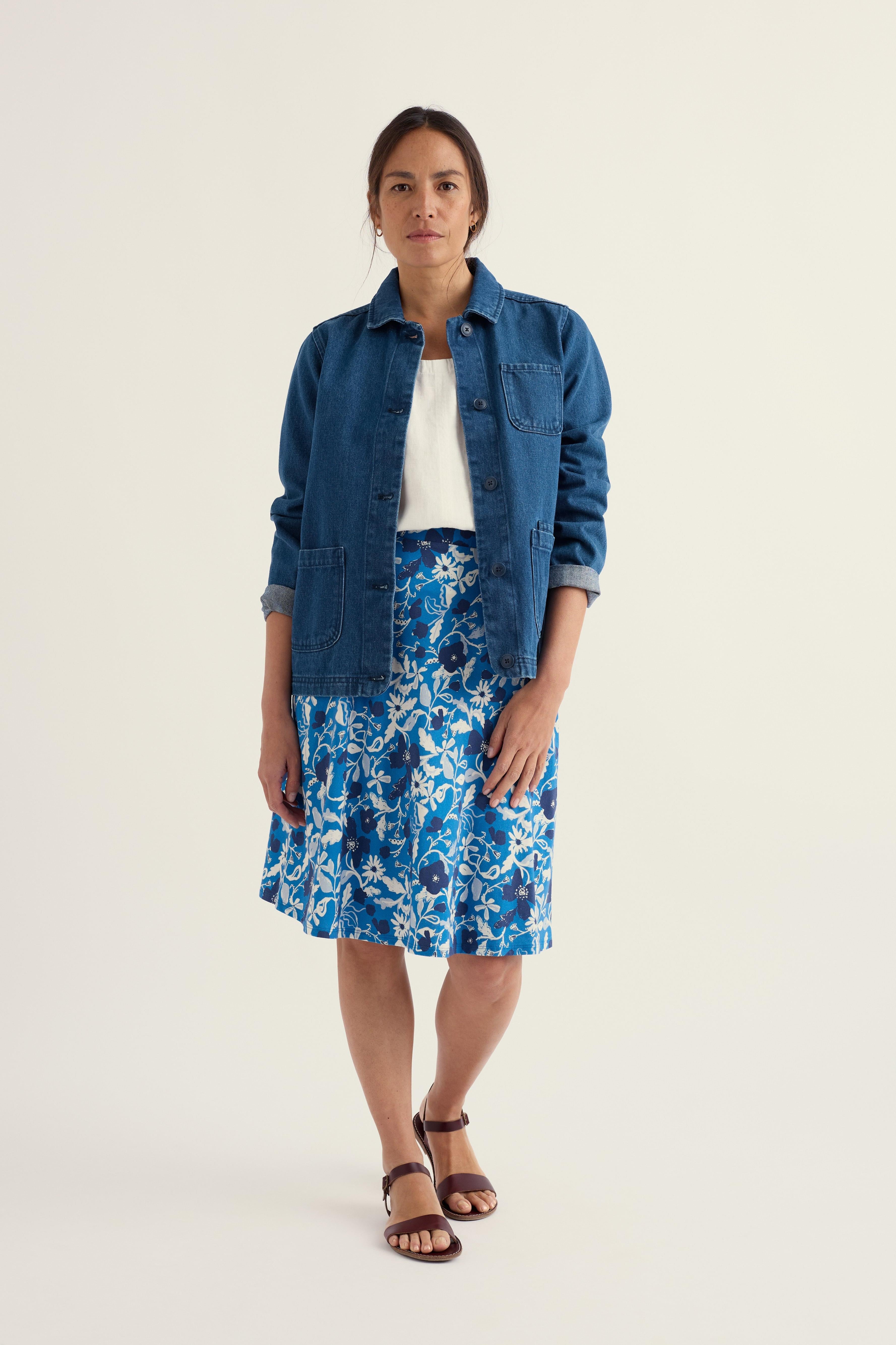Woman wearing a blue denim jacket over a white top and blue floral midi skirt against a plain background.