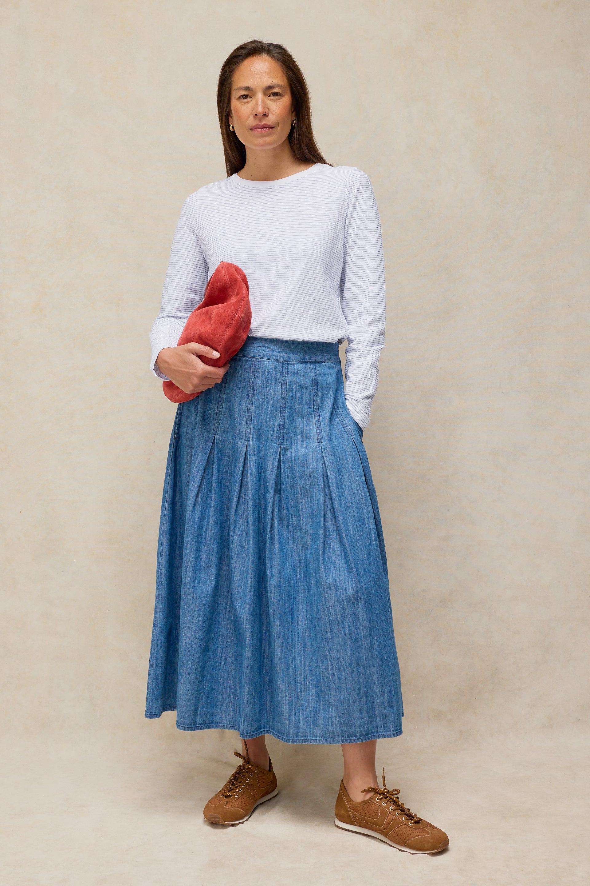 A woman wearing a striped cotton top and pleated denim skirt holding a red clutch against a neutral background.
