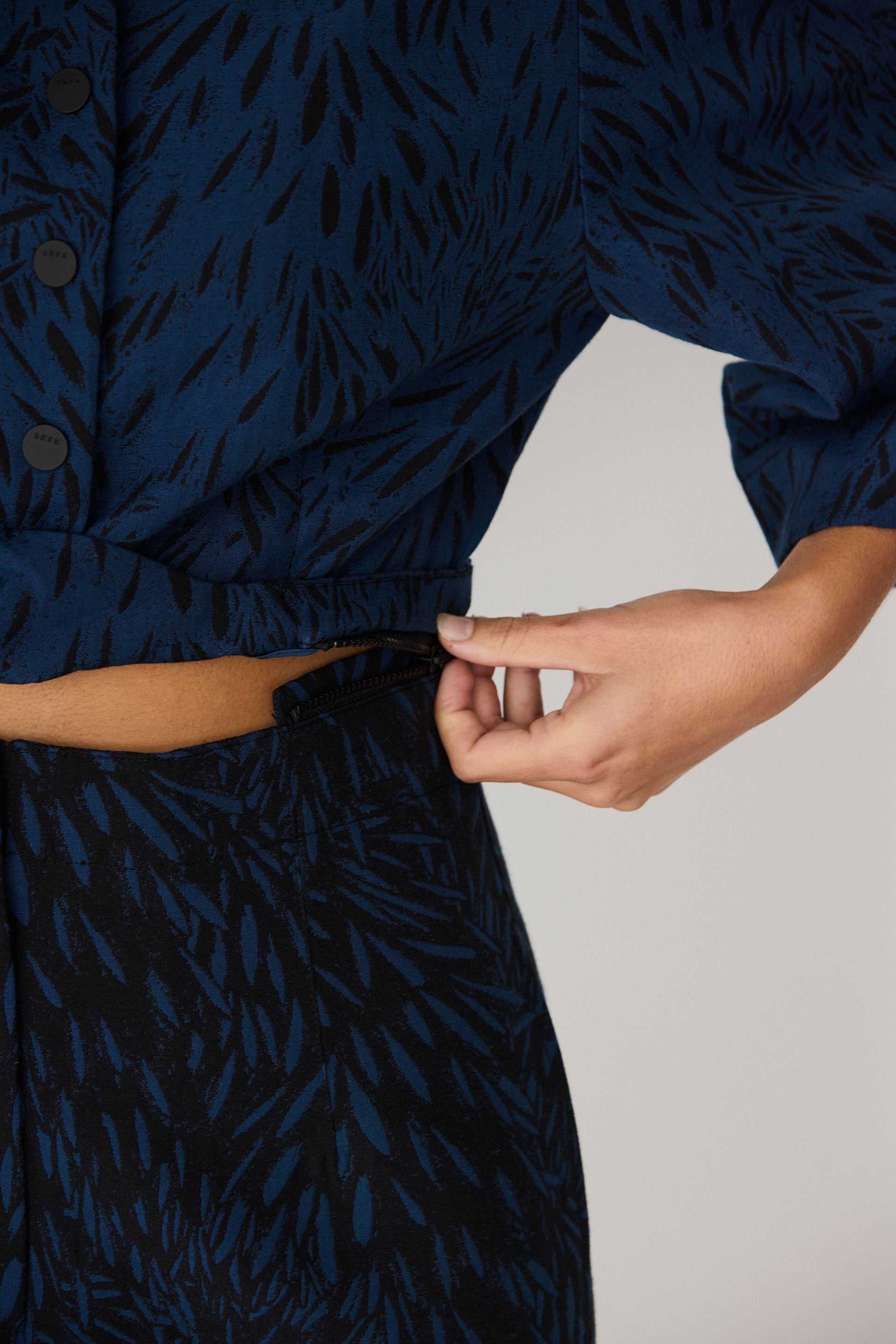Woman’s hand zipping or unzipping the waist of a blue patterned sustainable jumpsuit.