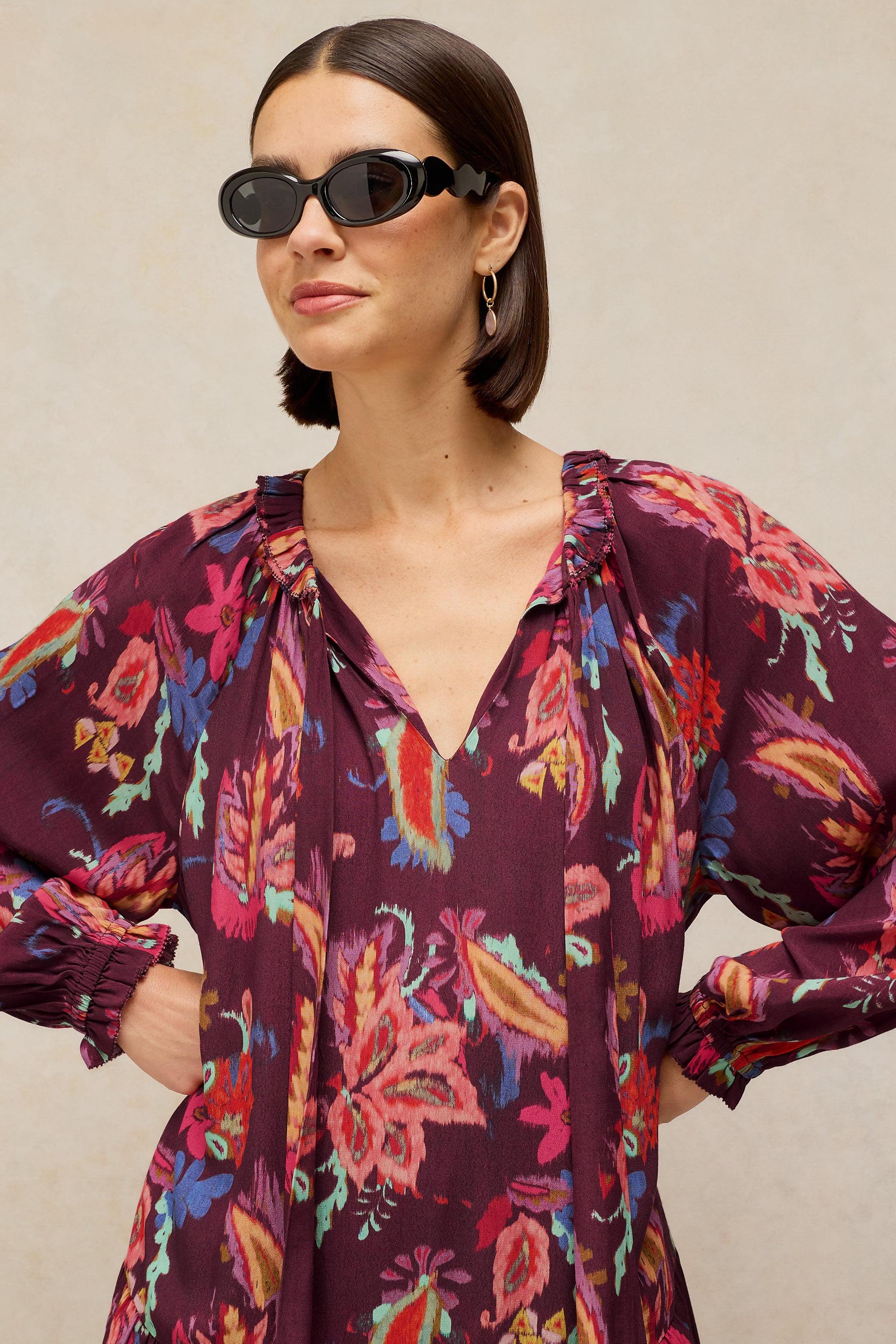 A woman wearing a long-sleeve, deep V-neck blouse with a colorful floral print on a dark purple background, styled with black sunglasses and gold hoop earrings against a neutral beige background.