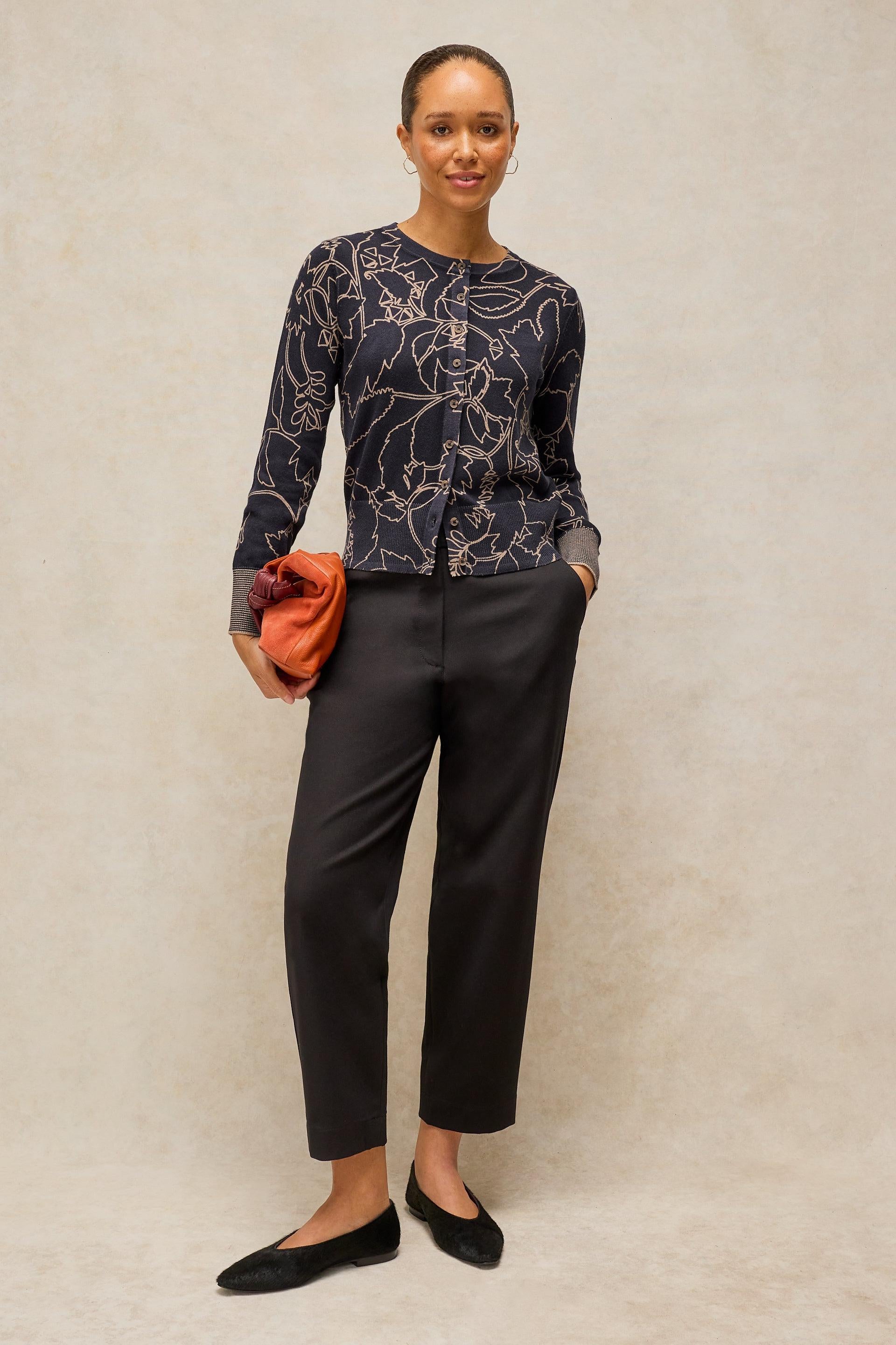 A woman modeling a dark navy long-sleeve top with a white abstract floral pattern, paired with black cropped trousers and black flats, holding a small orange clutch against a textured beige studio backdrop.