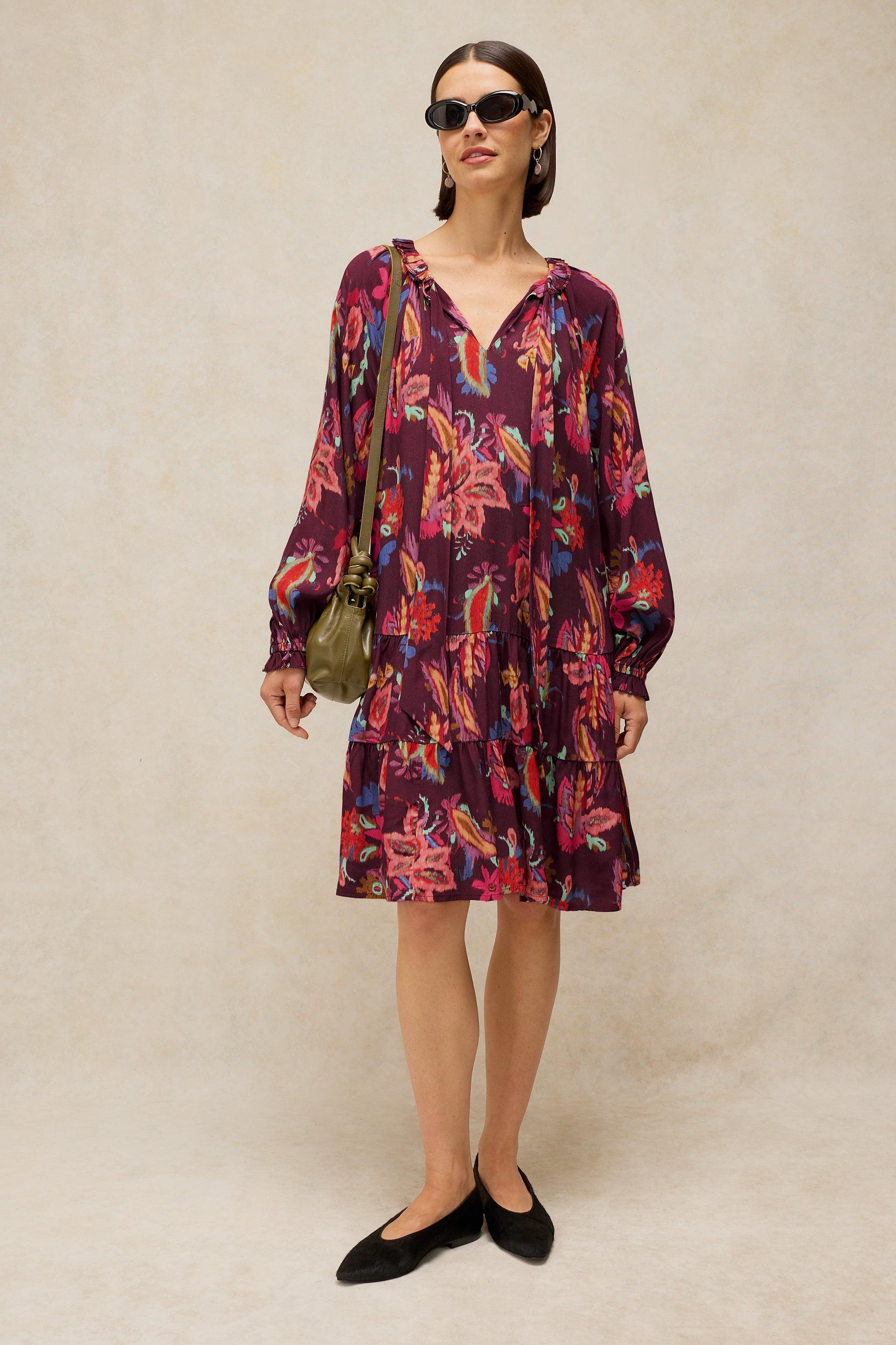 A woman modeling a long-sleeve, knee-length V-neck dress with a vibrant multicolor floral print on a dark purple base. She is wearing black sunglasses, a small olive green shoulder bag, and black flats, standing against a textured beige studio backdrop.
