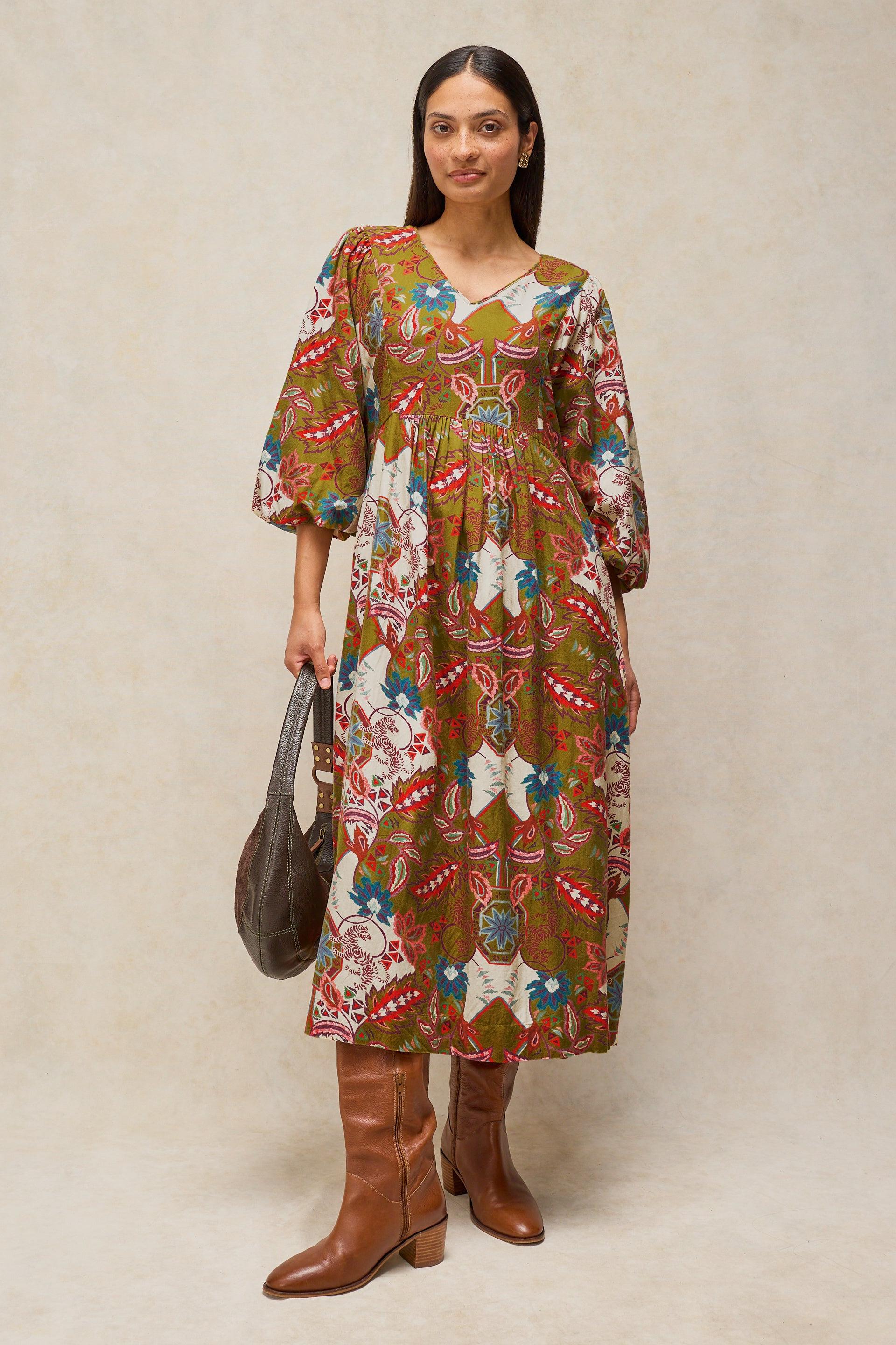 A woman modeling a bohemian-style midi dress with a multicolor abstract print on a brown base, paired with brown leather knee-high boots and holding a dark brown leather handbag, set against a neutral beige studio backdrop.
