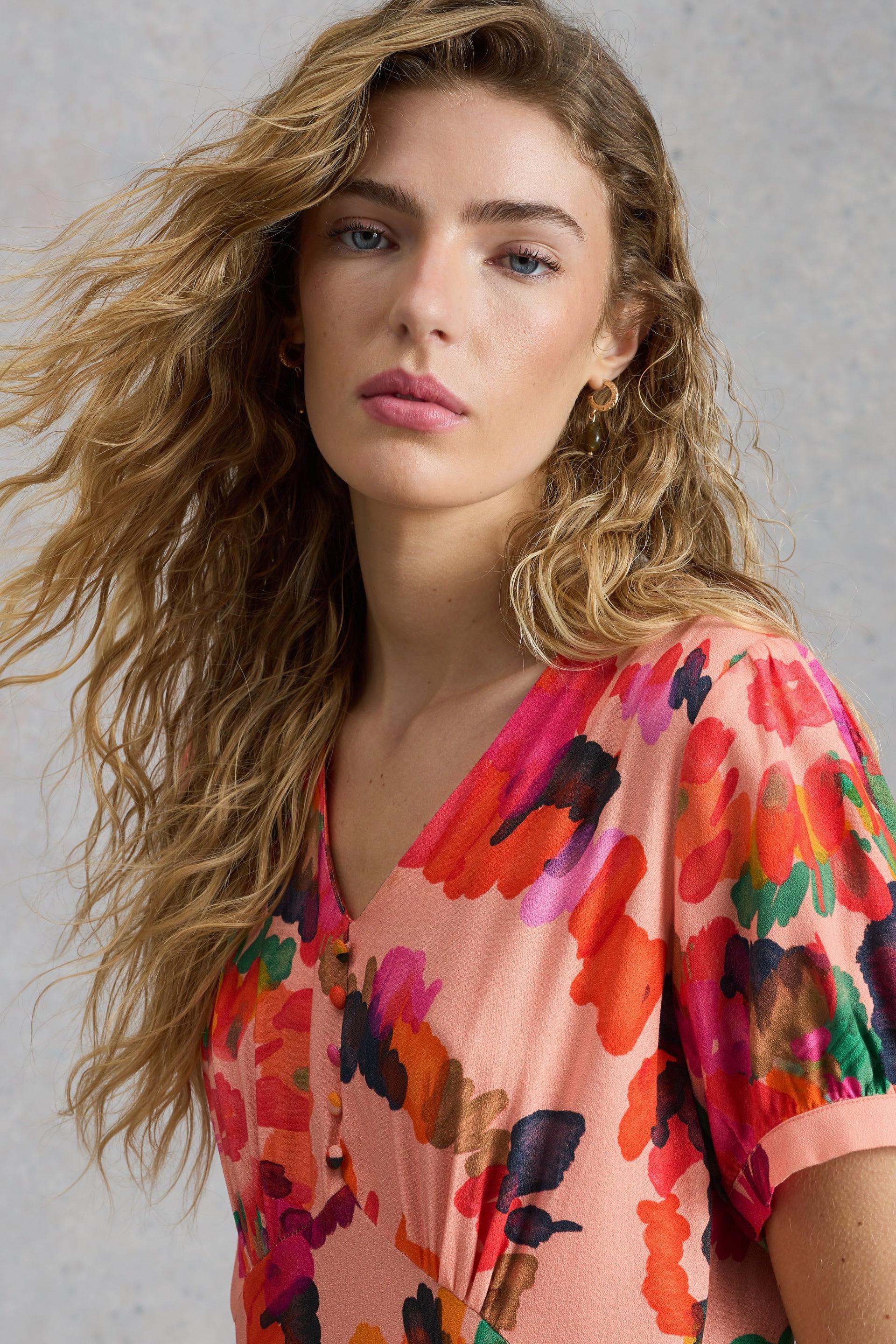 Woman with long wavy hair wearing a bright floral occasion dress against a neutral grey wall.
