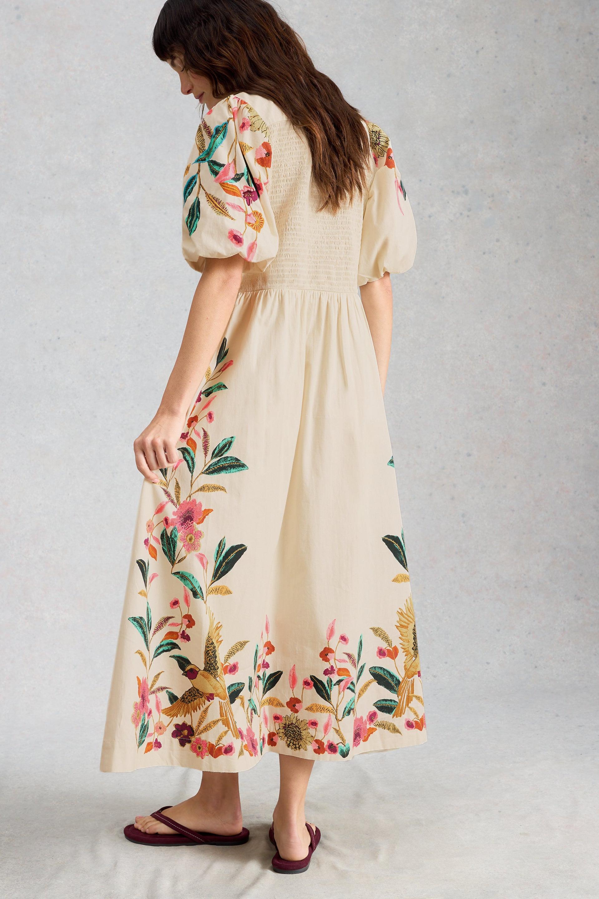 Woman wearing a beige organic cotton occasion dress with vibrant floral embroidery and puff sleeves.