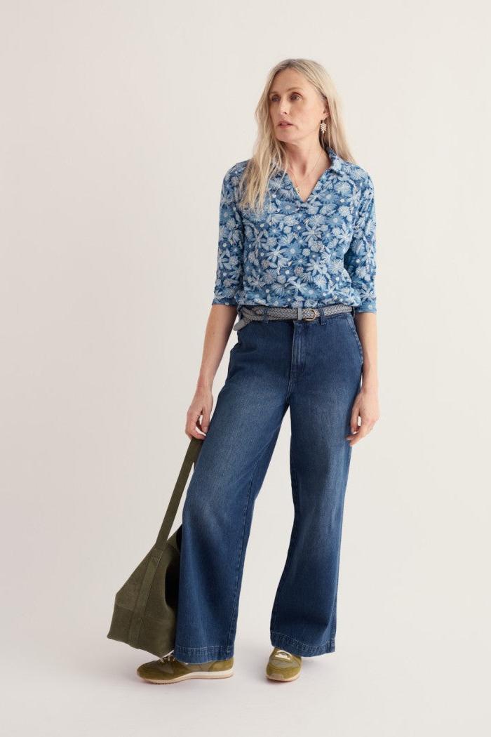 A woman modeling a blue floral button-up shirt, dark blue wide-leg jeans, olive green shoes, and carrying an olive green tote bag against a plain light beige studio background.
