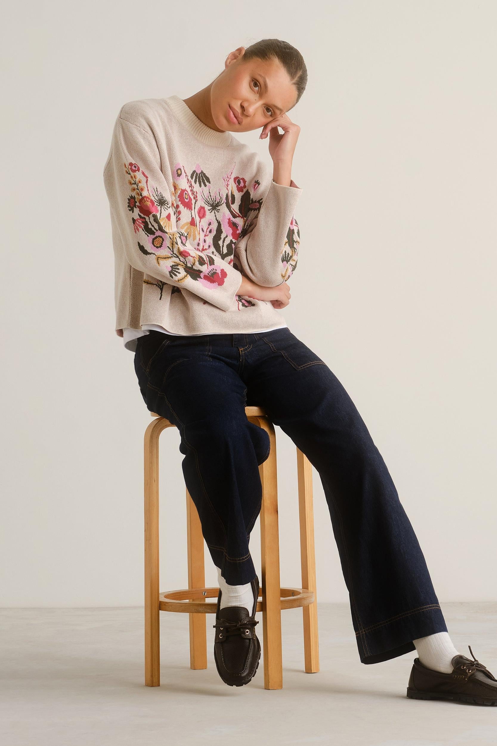 A model sits on a wooden stool wearing a cream knit sweater with colorful floral embroidery on the sleeves, dark wide-leg jeans, white socks, and dark brown loafers against a plain off-white studio background.