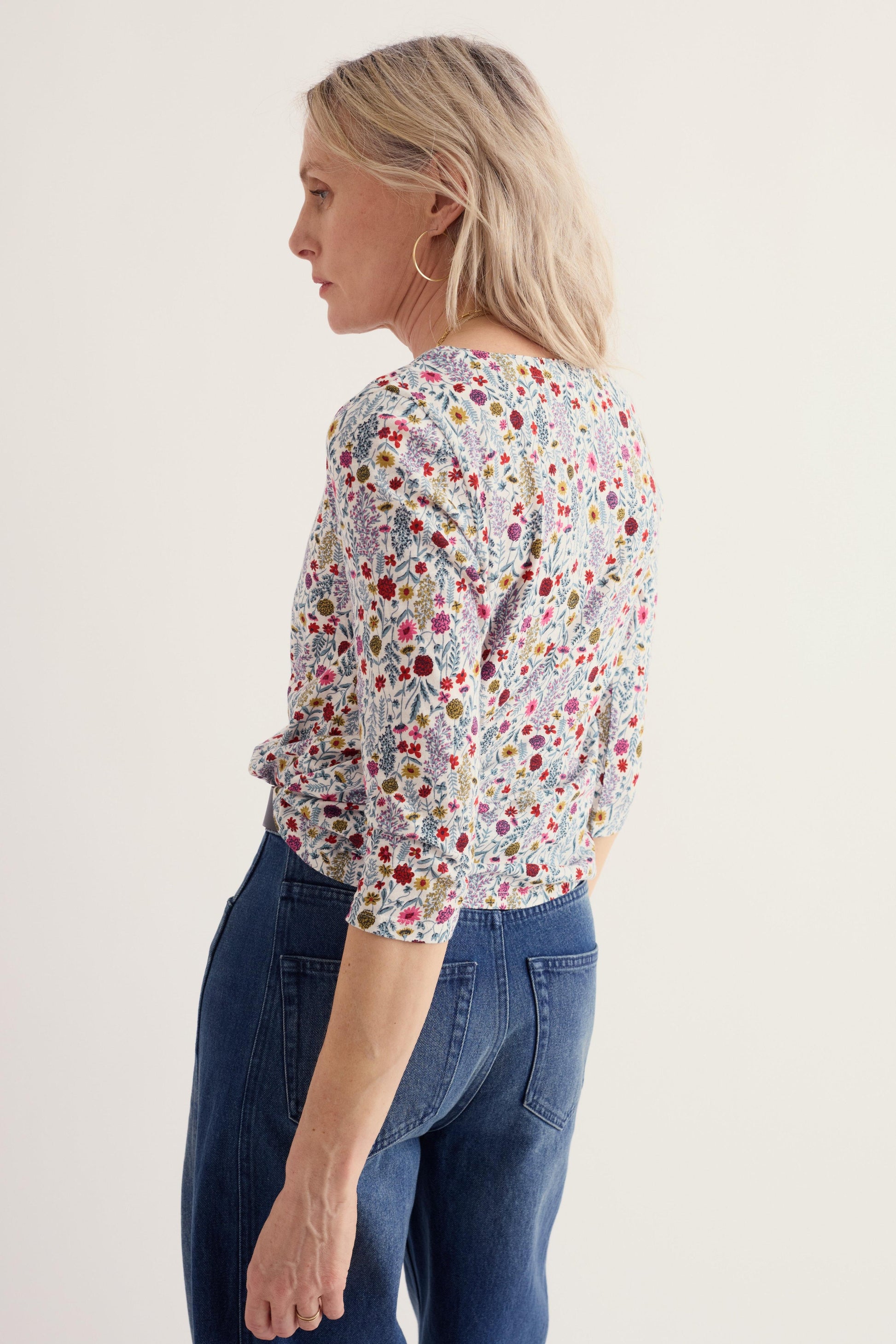 A woman shown from the back wearing a short-sleeve blouse with a dense multicolor floral and paisley print on a light background, paired with dark blue jeans, against a plain off-white studio background.