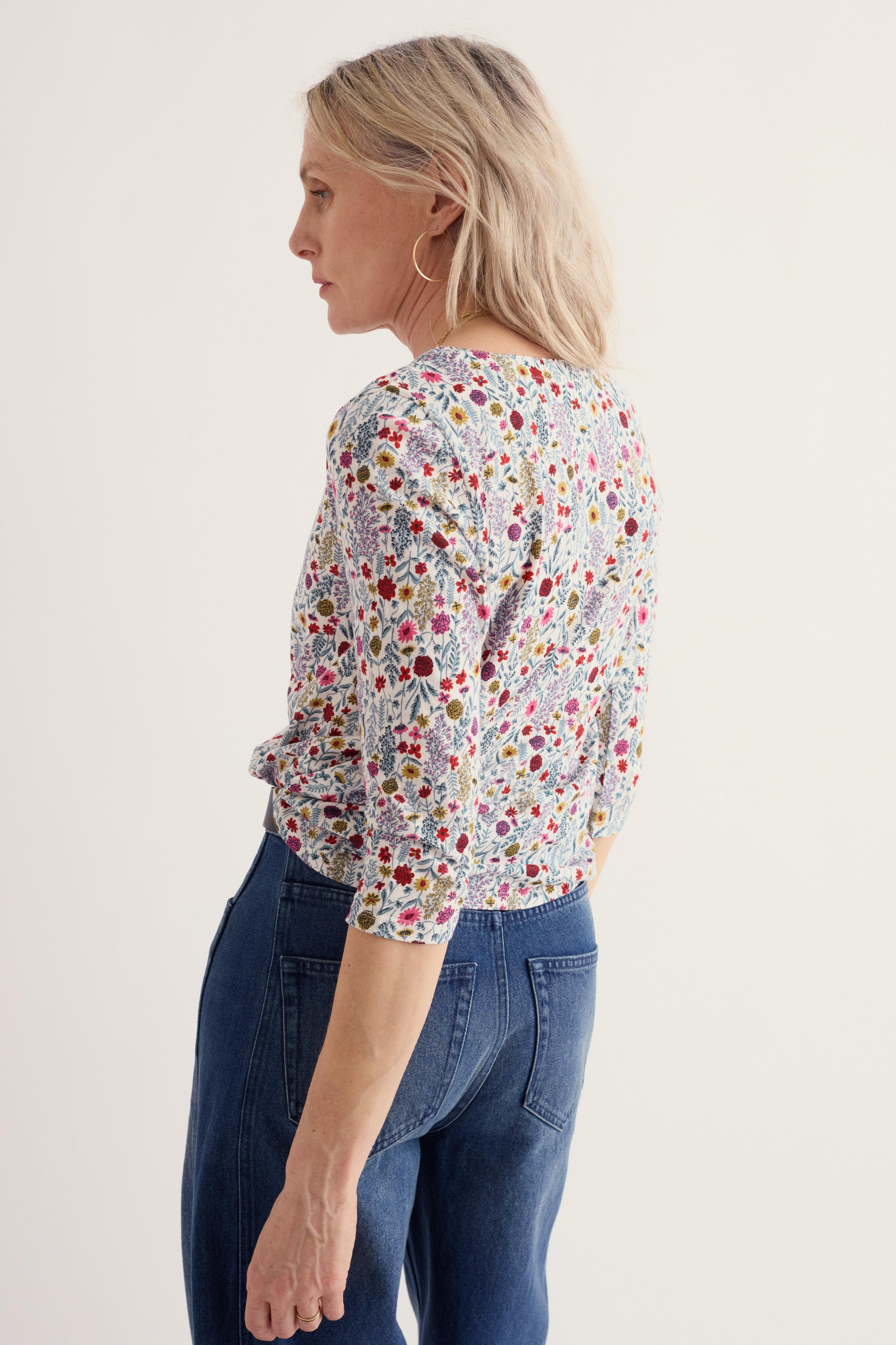 A woman shown from the back wearing a short-sleeve blouse with a dense multicolor floral and paisley print on a light background, paired with dark blue jeans, against a plain off-white studio background.