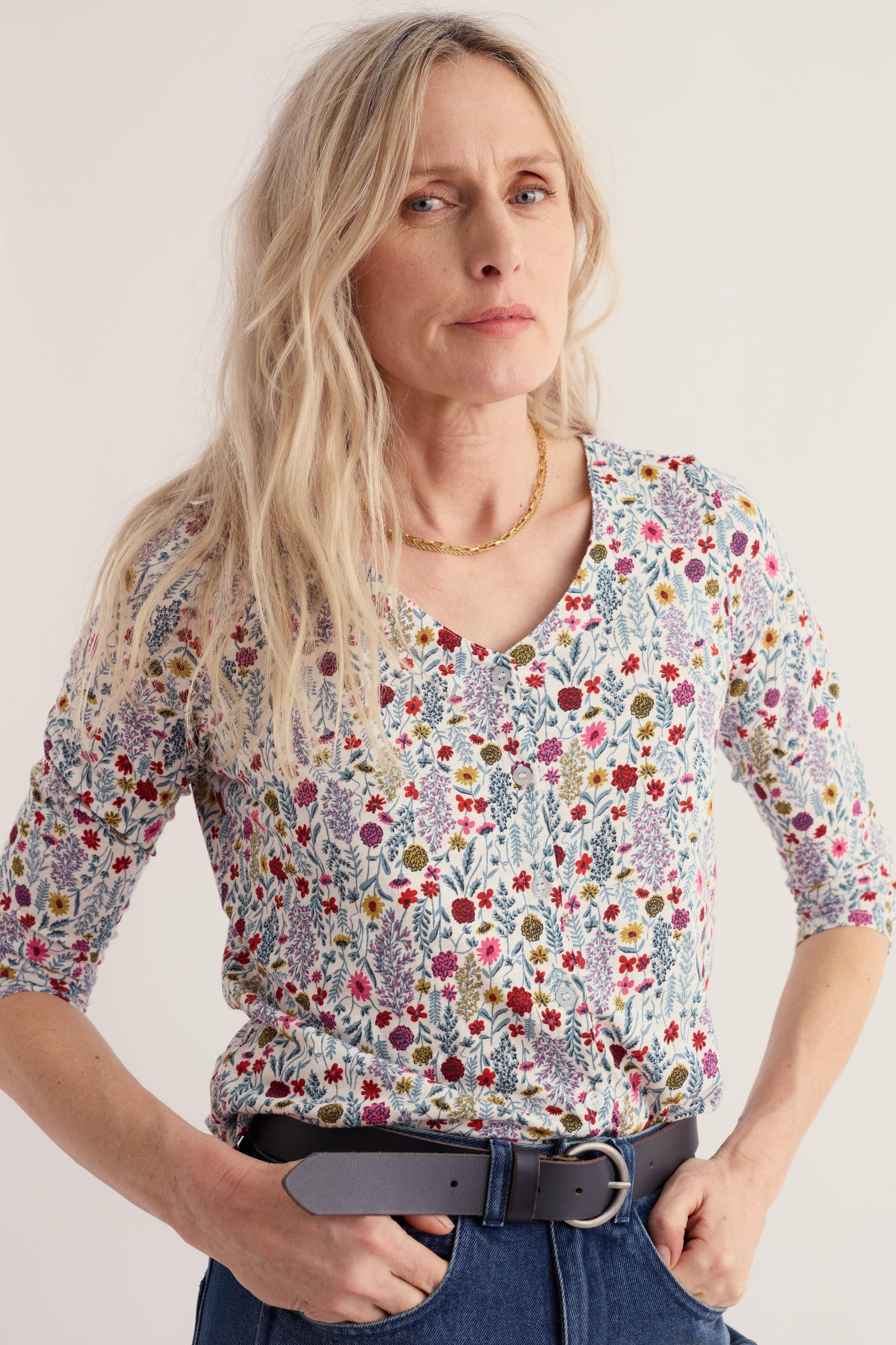 A woman wearing a short-sleeve V-neck blouse with a dense multicolor floral and botanical print on a light background, paired with a black belt and blue jeans, against a plain light background.