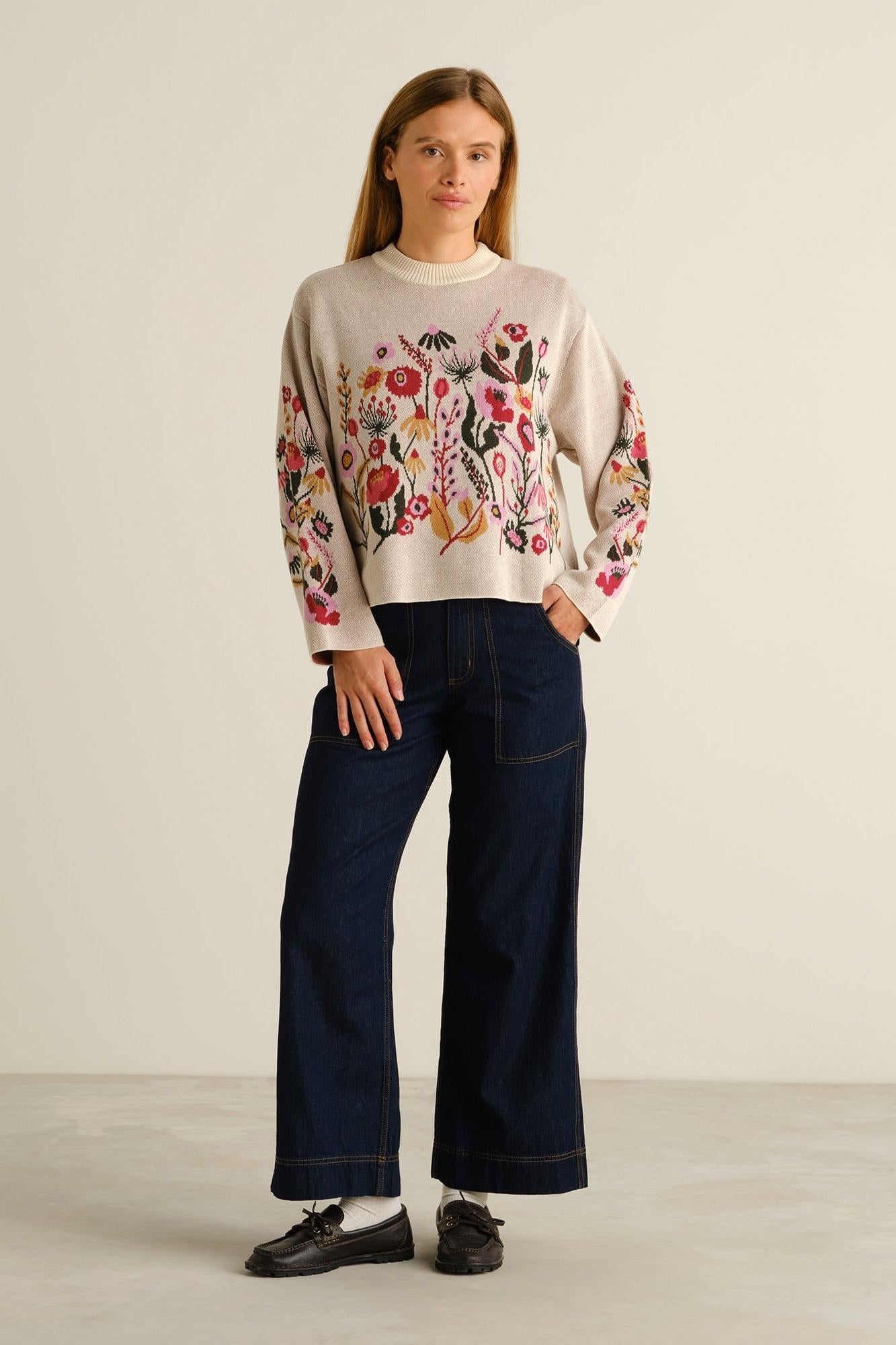 A woman stands in a studio wearing a cream knit sweater with colorful floral embroidery on the front and sleeves, paired with dark wide-leg jeans and black loafers.