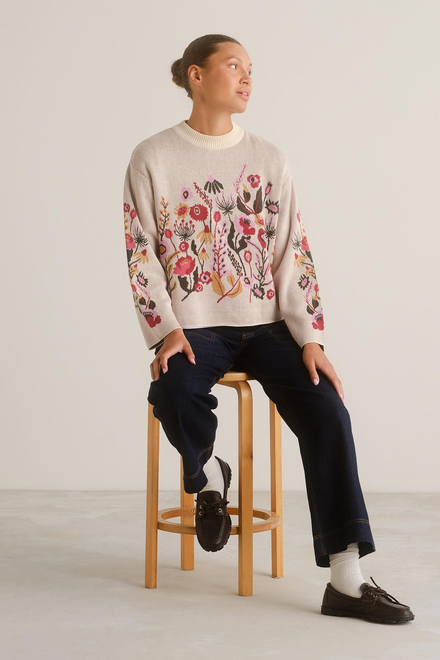 A model sits on a wooden stool wearing a cream-colored crewneck sweater with colorful floral embroidery on the front and sleeves, paired with dark blue jeans, white socks, and black loafers.