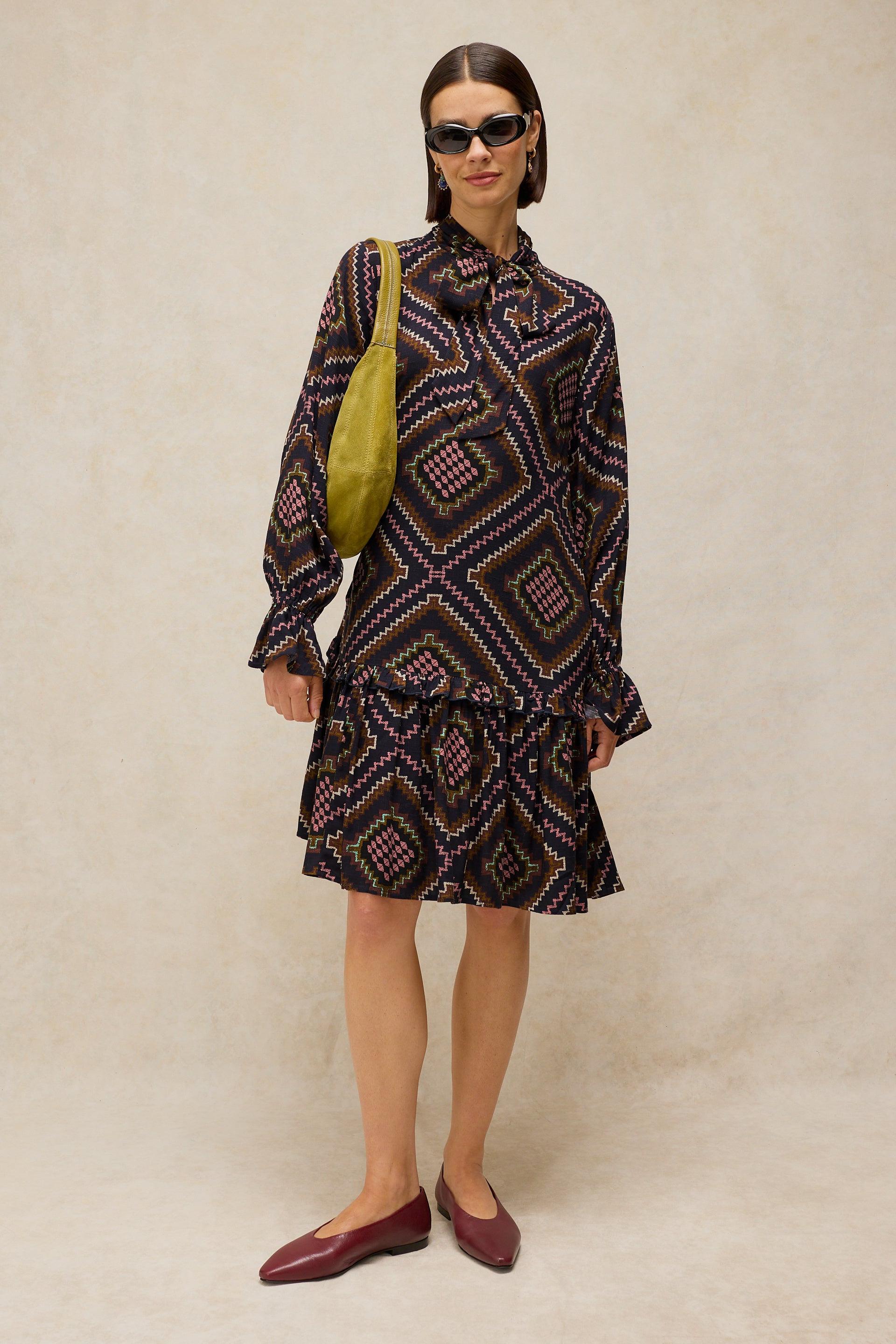 Woman wearing a patterned dress with a green bag against a beige background