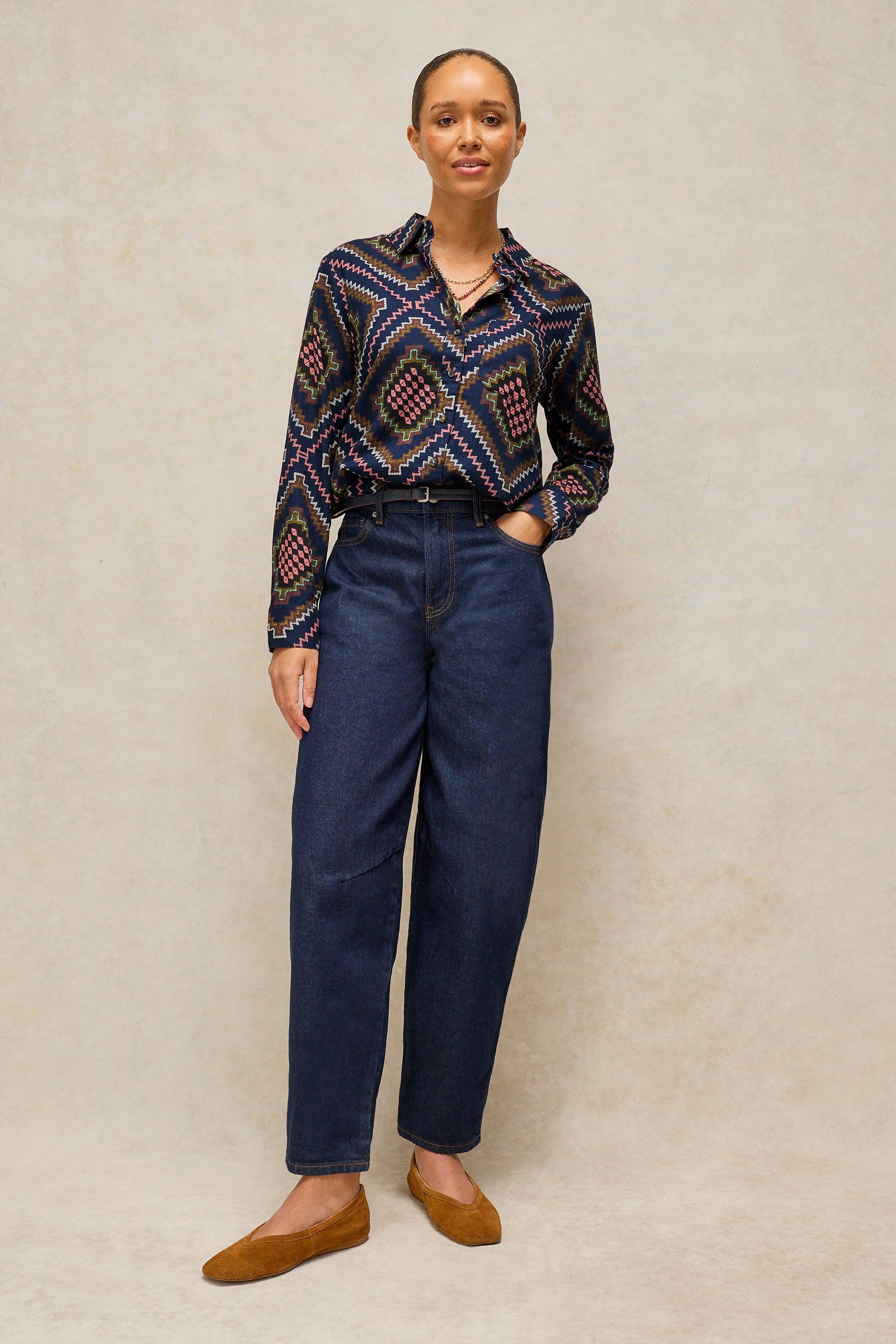 A woman stands against a textured beige studio backdrop wearing a long-sleeve button-up shirt with a dark navy and red geometric diamond pattern, dark blue jeans, and tan flats.