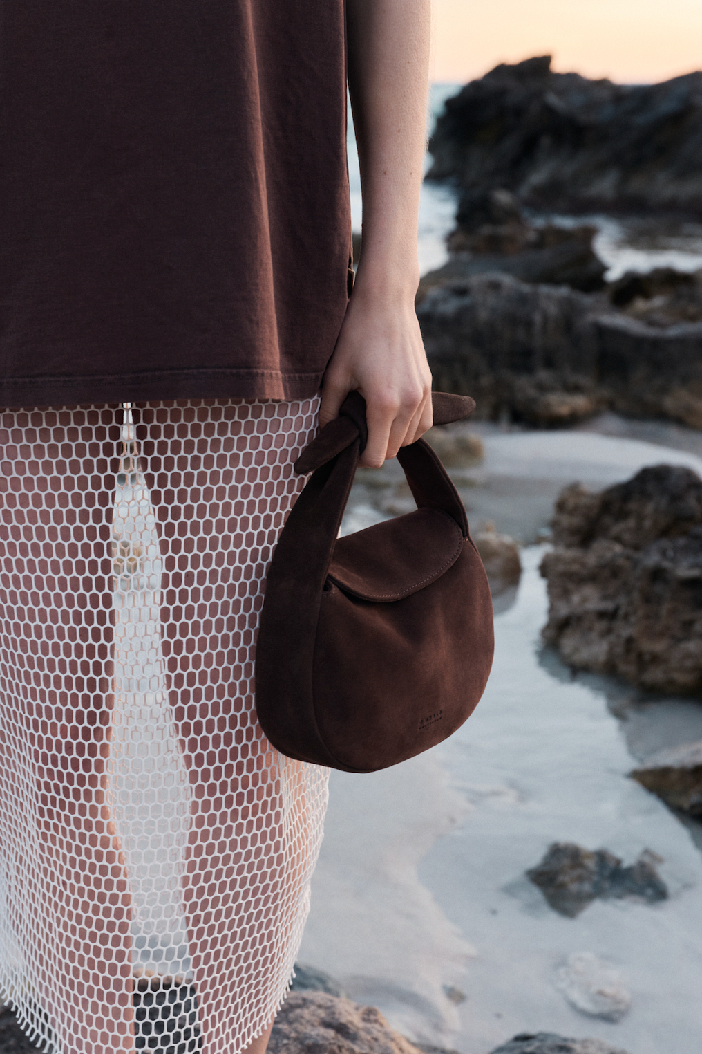 Person holding a brown suede handbag on a rocky beach