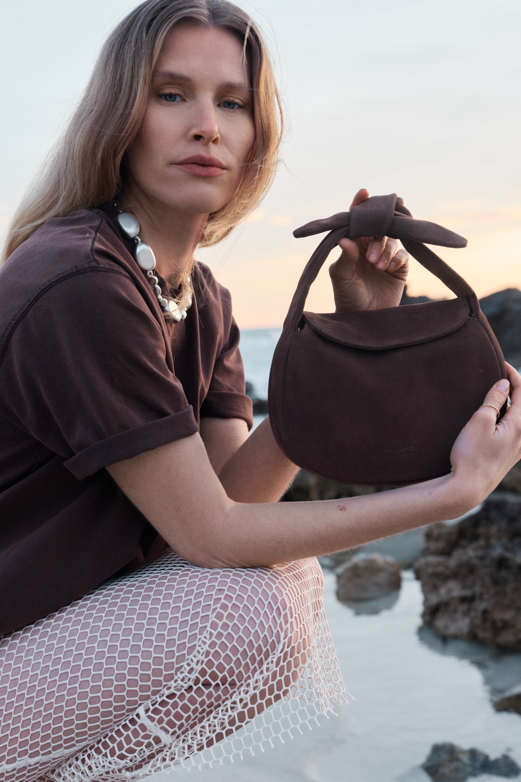 Woman holding a brown handbag by the water during sunset