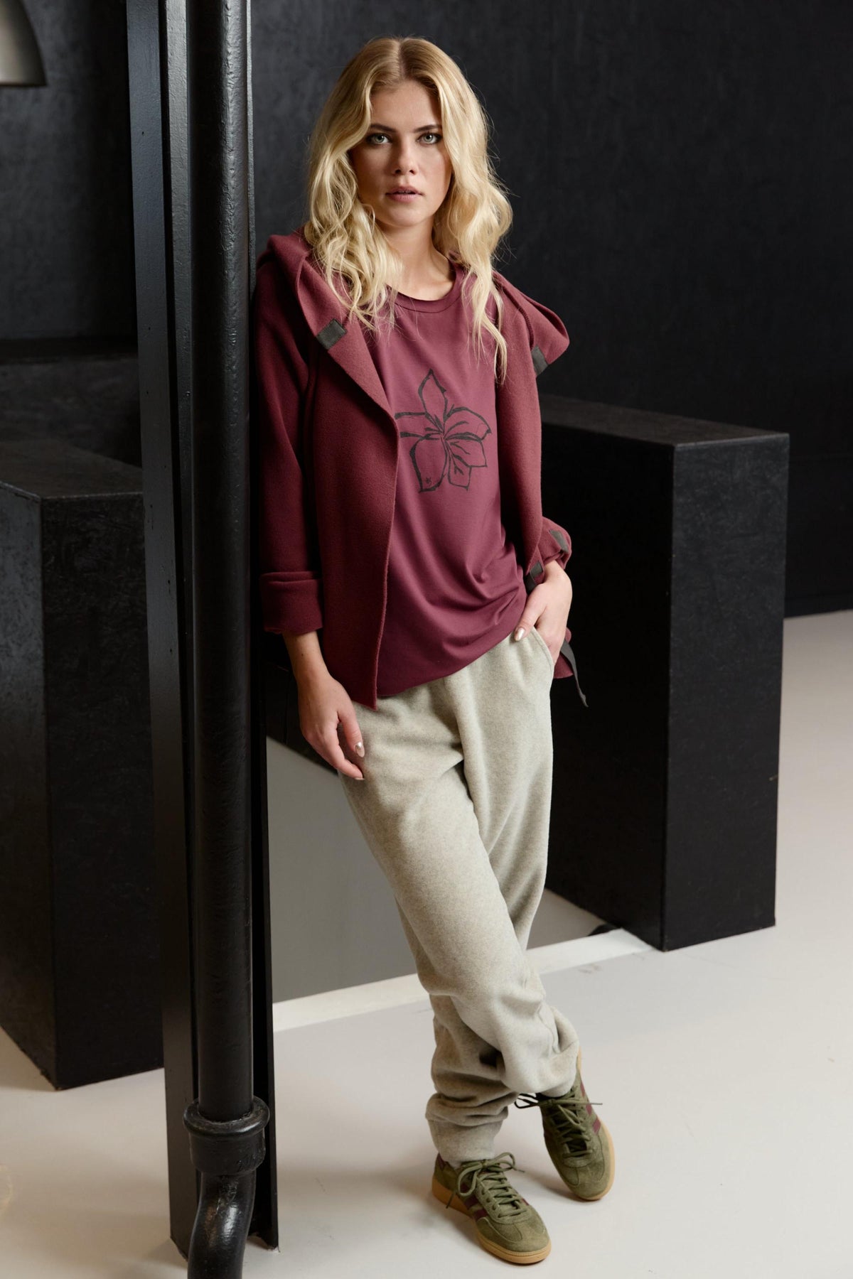A woman wearing a burgundy Henriette Steffensen fleece cardigan and light grey trousers leaning against a black pillar.