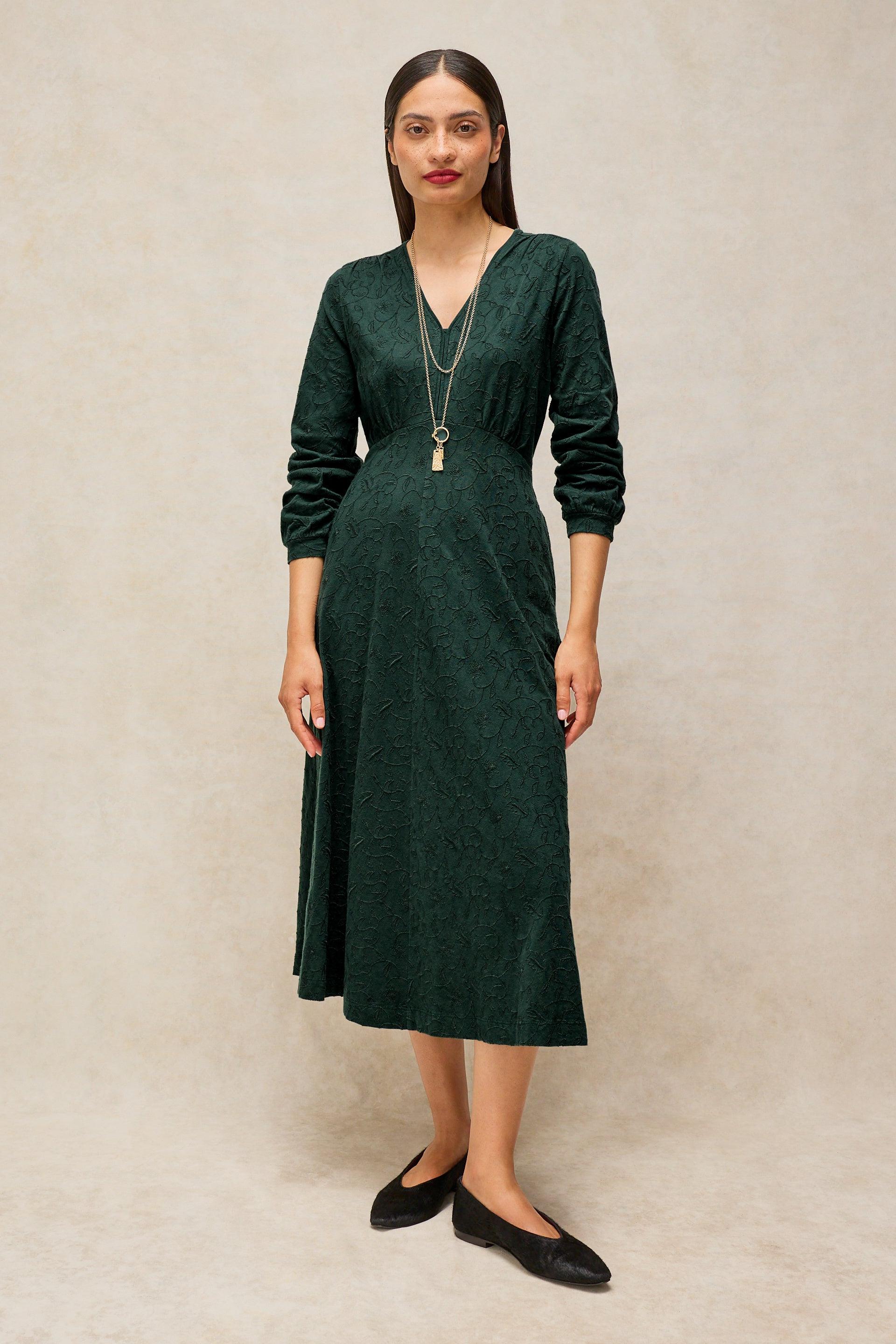 A woman modeling a dark green long-sleeve midi dress with a V-neckline, standing against a textured beige studio backdrop.