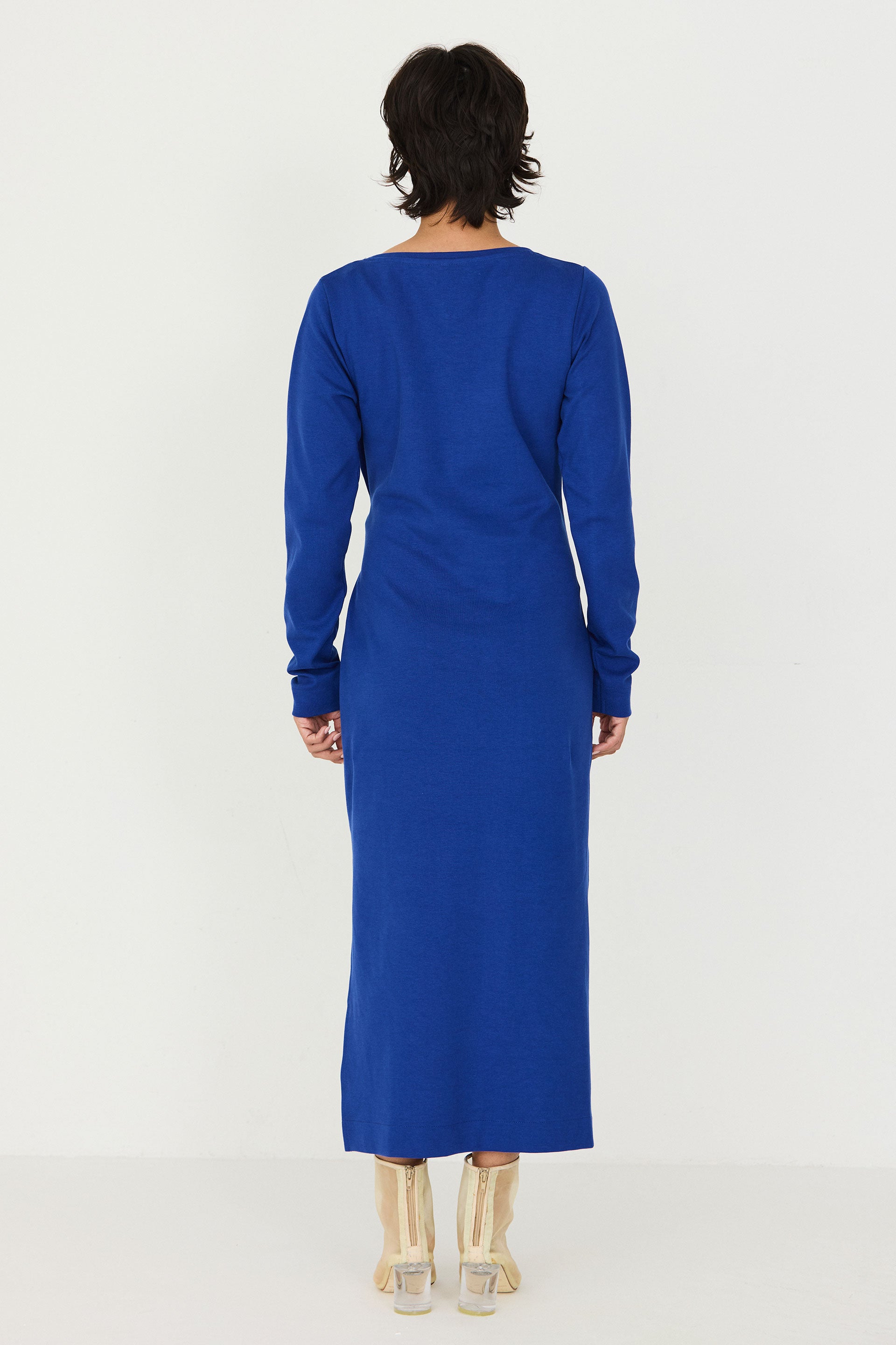 A woman viewed from the back wearing a long-sleeve royal blue midi dress and beige high-heeled shoes against a plain white studio background.