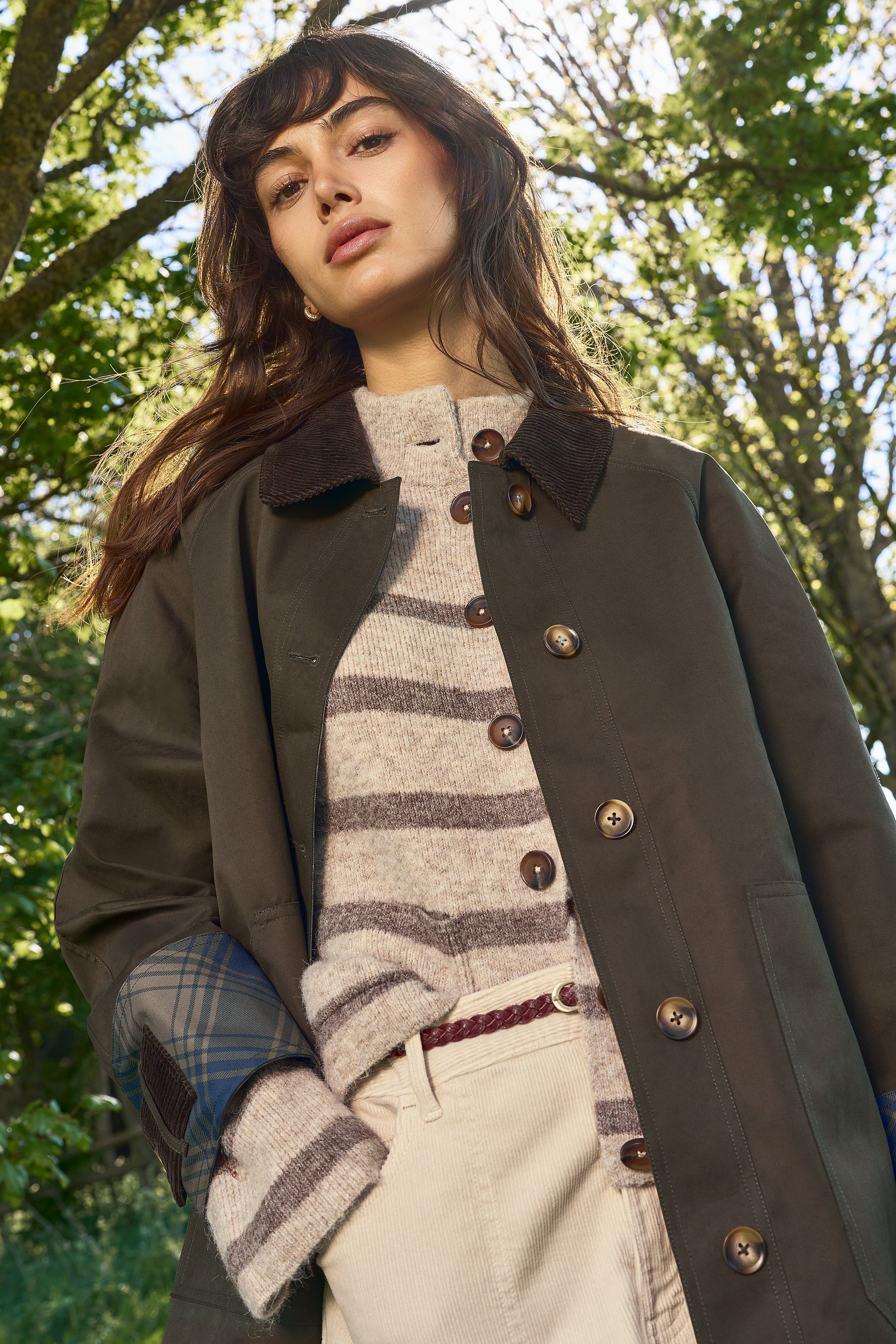 A woman wearing a dark brown wool-blend coat over a striped knit sweater and cream trousers, standing outdoors in a sunlit wooded setting.