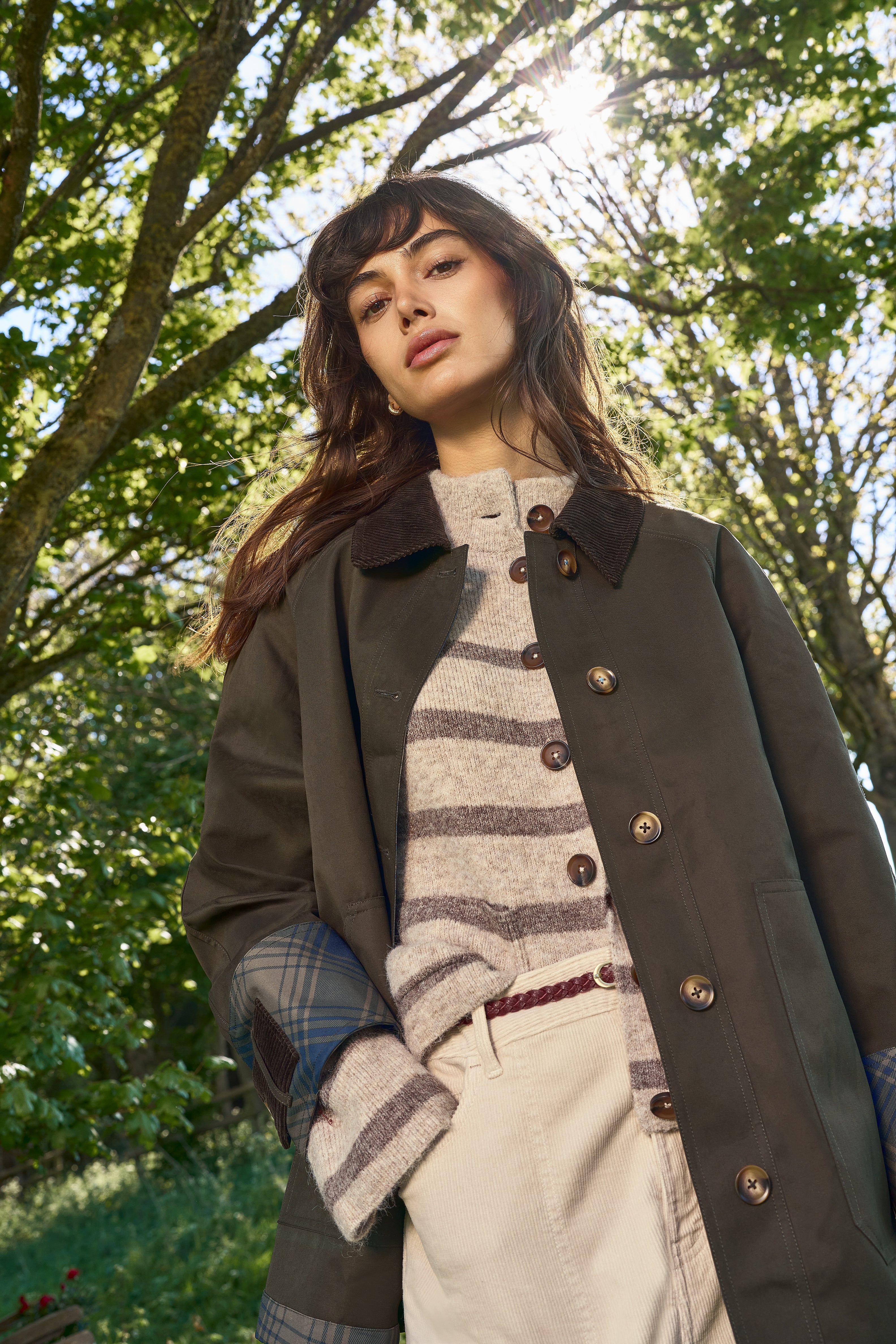 A woman wearing a dark brown wool-blend coat over a striped knit sweater and cream trousers, standing outdoors in a sunlit wooded setting.