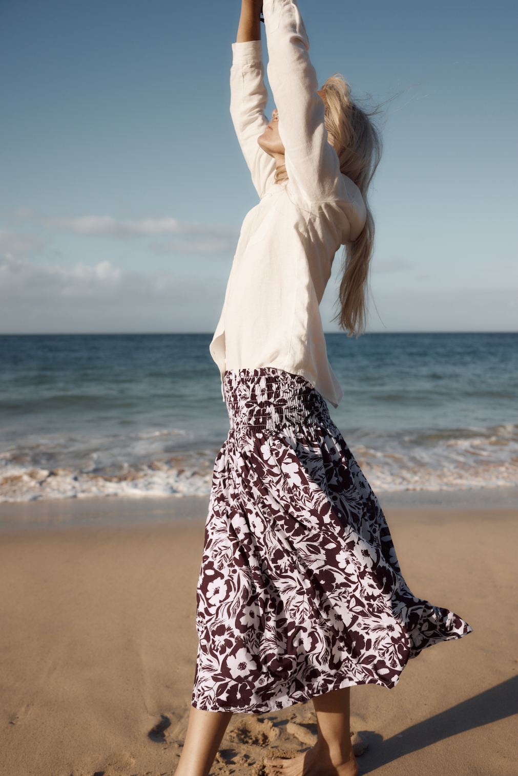A woman wearing a Seasalt organic cotton floral skirt stretching her arms on a sunny beach.