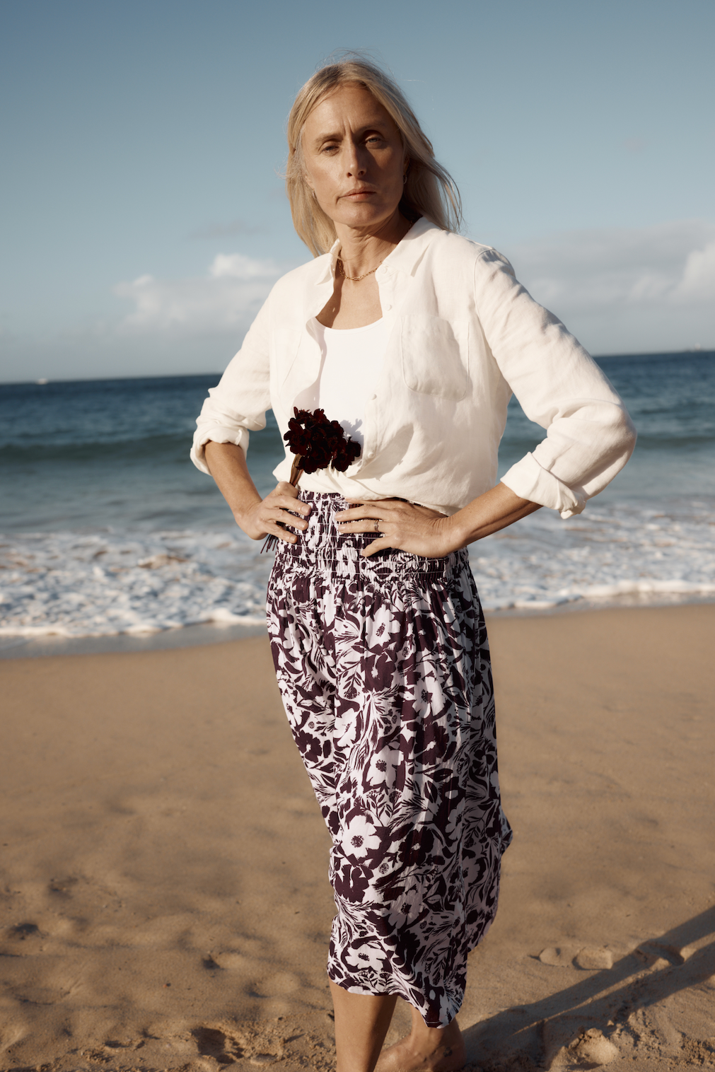 Blonde woman wearing a Seasalt organic cotton floral skirt and white shirt standing on a beach.
