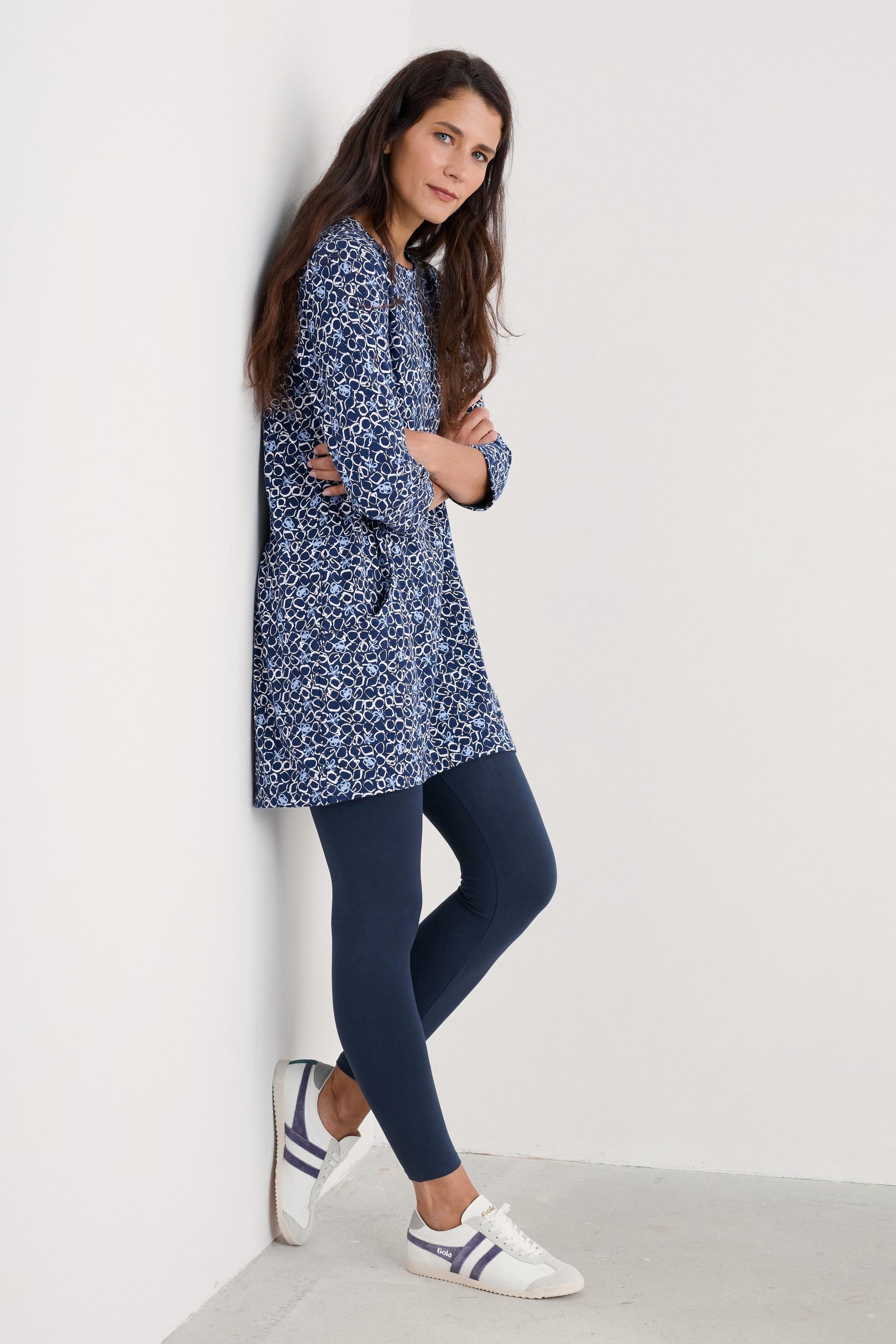 A woman leans against a white wall wearing a navy blue and white patterned long-sleeve tunic top, dark navy leggings, and white sneakers with navy stripes.