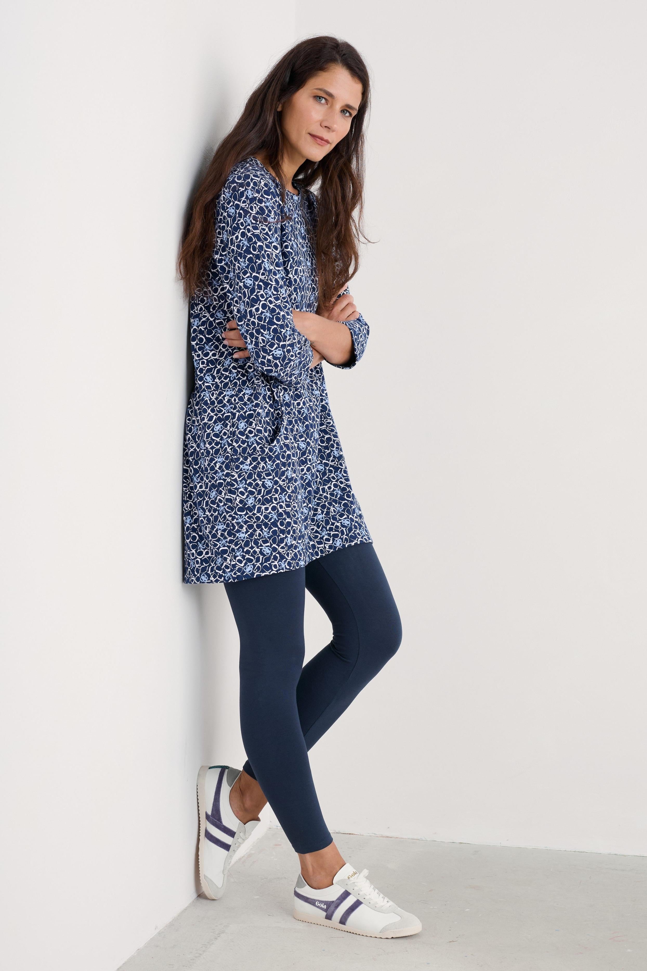 A woman leans against a white wall wearing a navy blue and white patterned long-sleeve tunic top, dark navy leggings, and white sneakers with navy stripes.