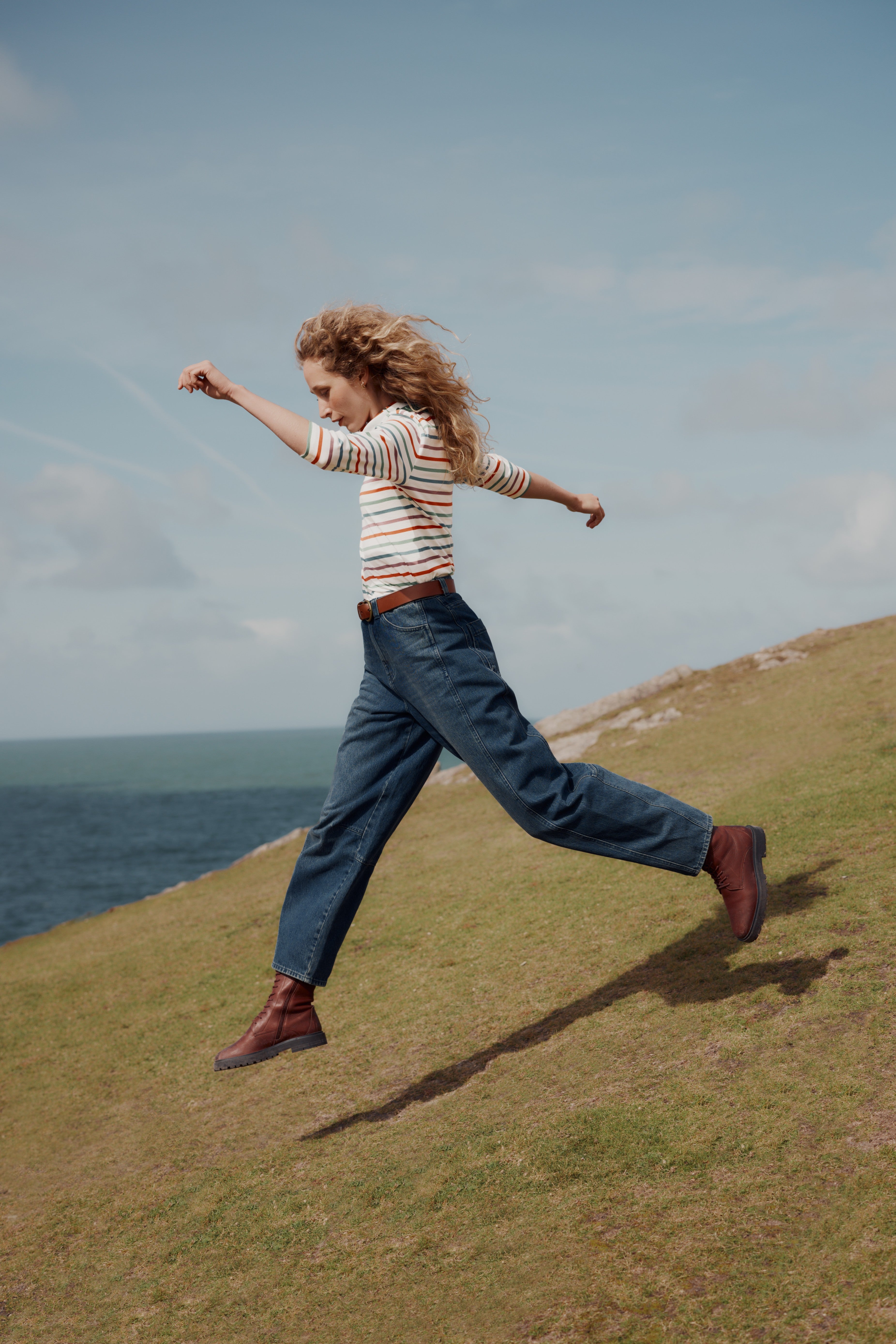 A woman with curly hair jumps across a grassy hill overlooking the ocean, wearing a horizontally striped long-sleeve top, loose dark blue jeans, and brown boots.