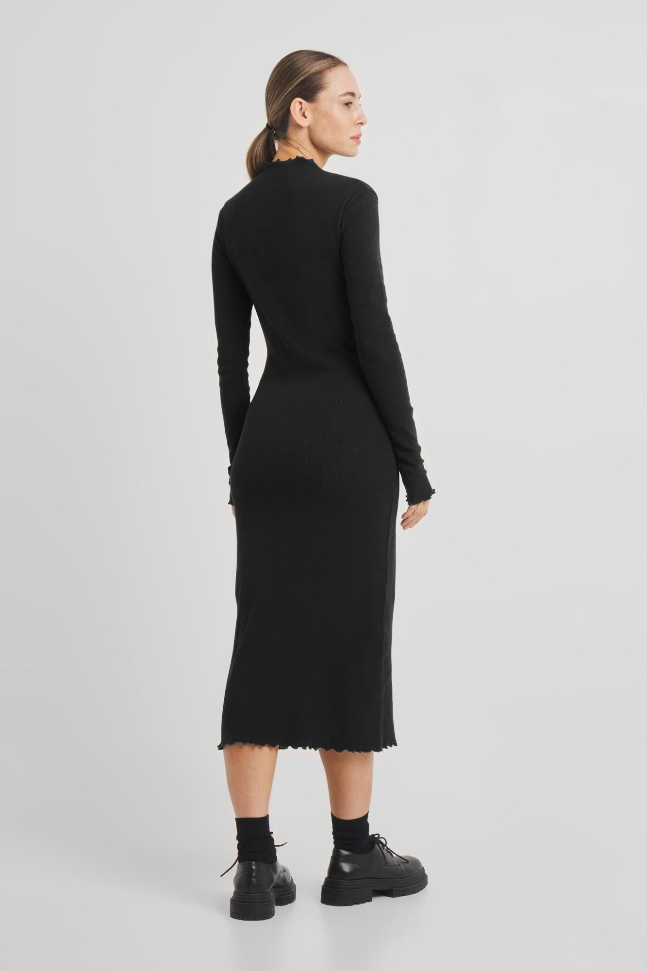 A woman modeling a black long-sleeve ribbed midi dress from the back, paired with black socks and black lace-up boots, against a light gray studio background.