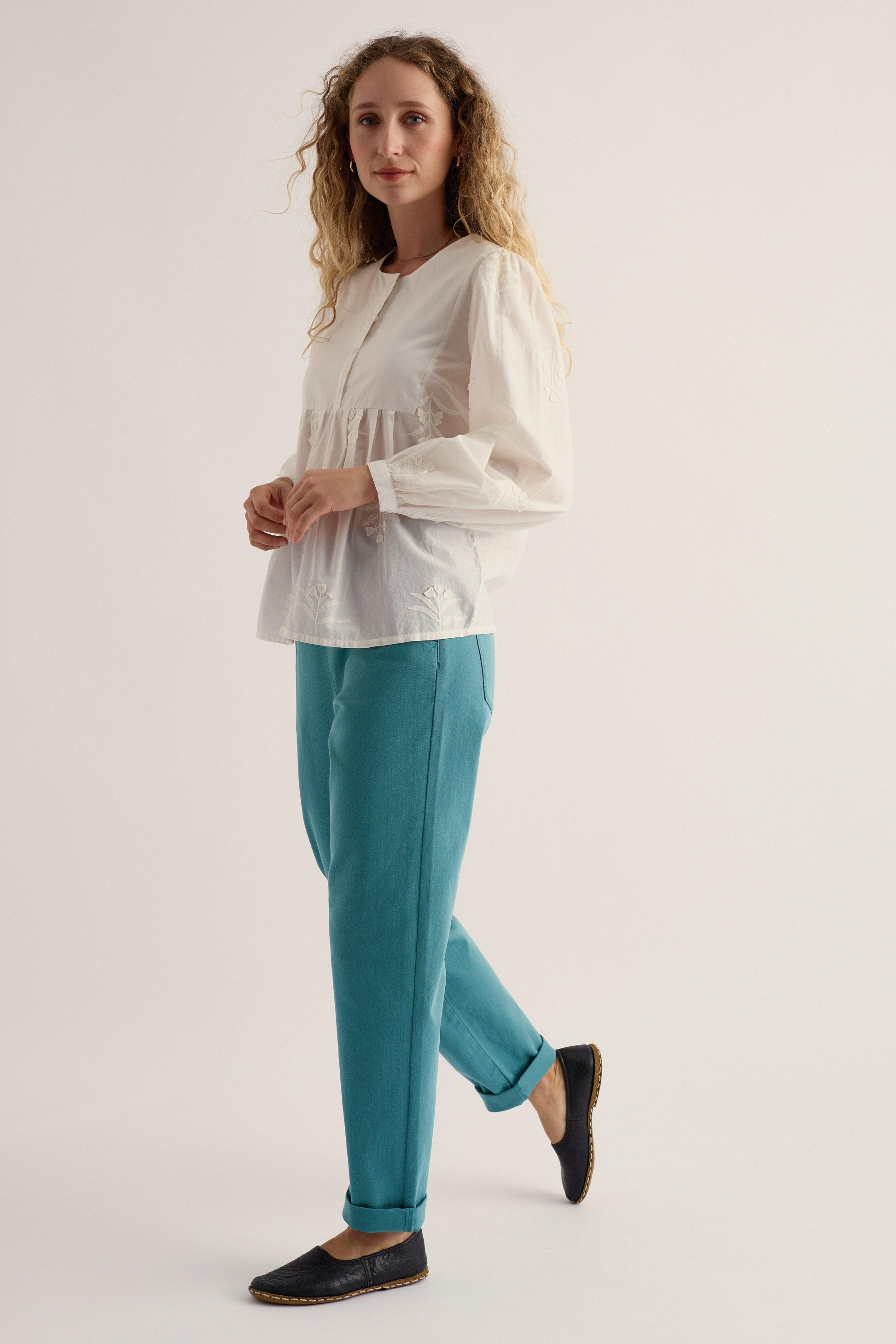 Woman wearing a white blouse and teal pants on a plain background