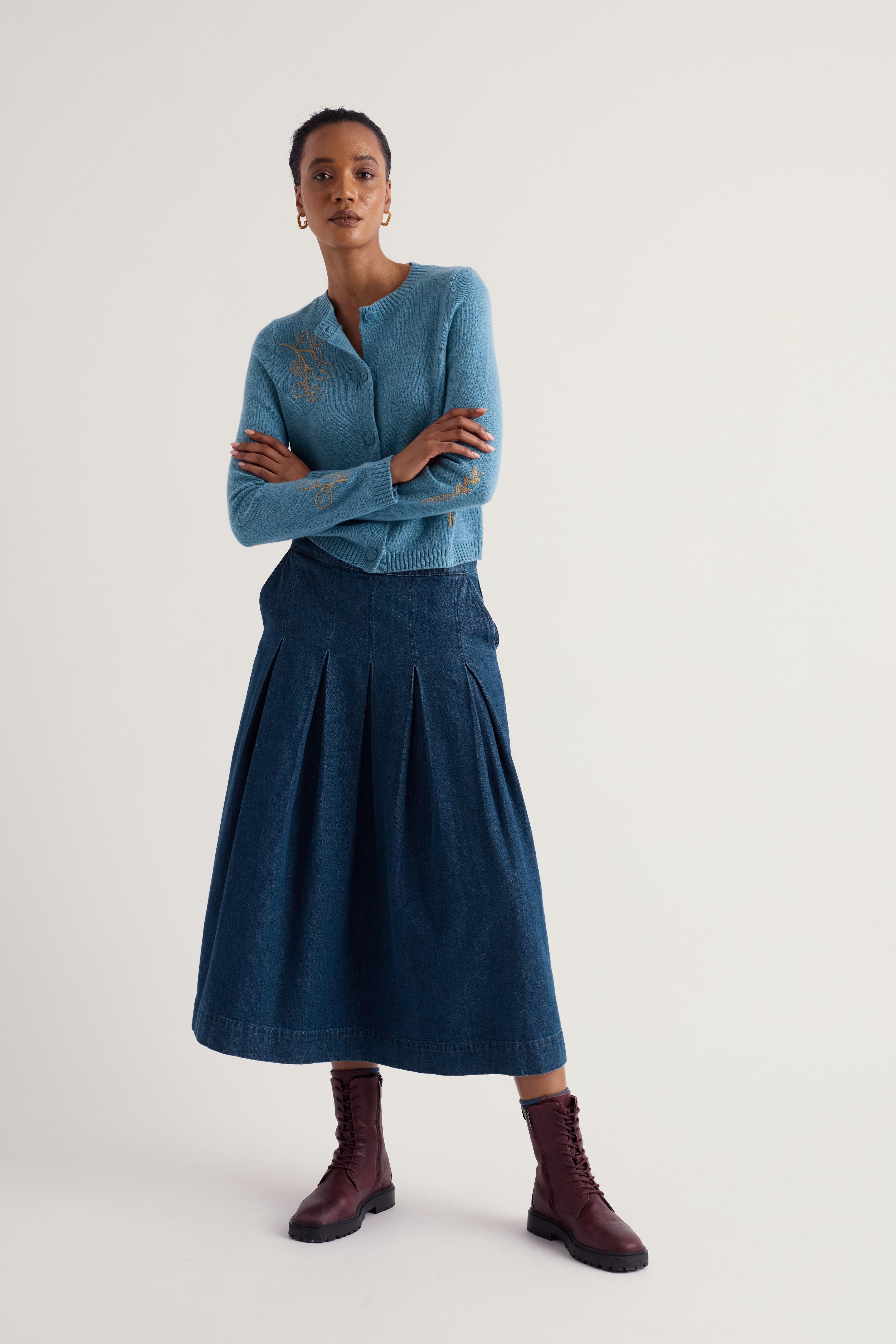 A model stands against a light gray studio background wearing a light blue long-sleeve knit top with subtle gold embroidery on the chest, a dark blue pleated midi skirt, and dark burgundy ankle boots.