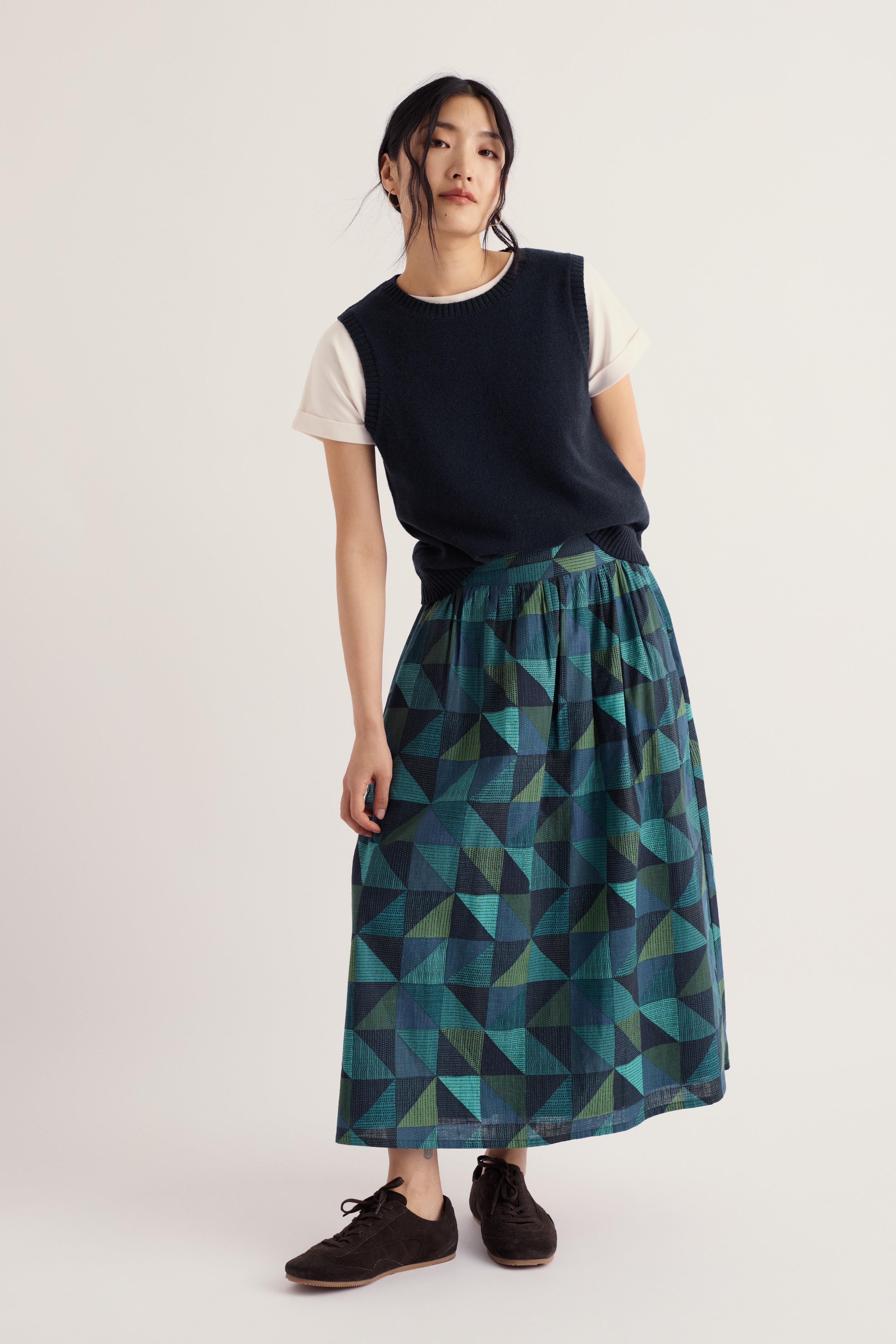 A model stands against a light off-white studio background wearing a sleeveless dark navy top over a white t-shirt and a pleated midi skirt with a geometric triangle pattern in teal, green, and dark blue. She is wearing dark brown shoes.