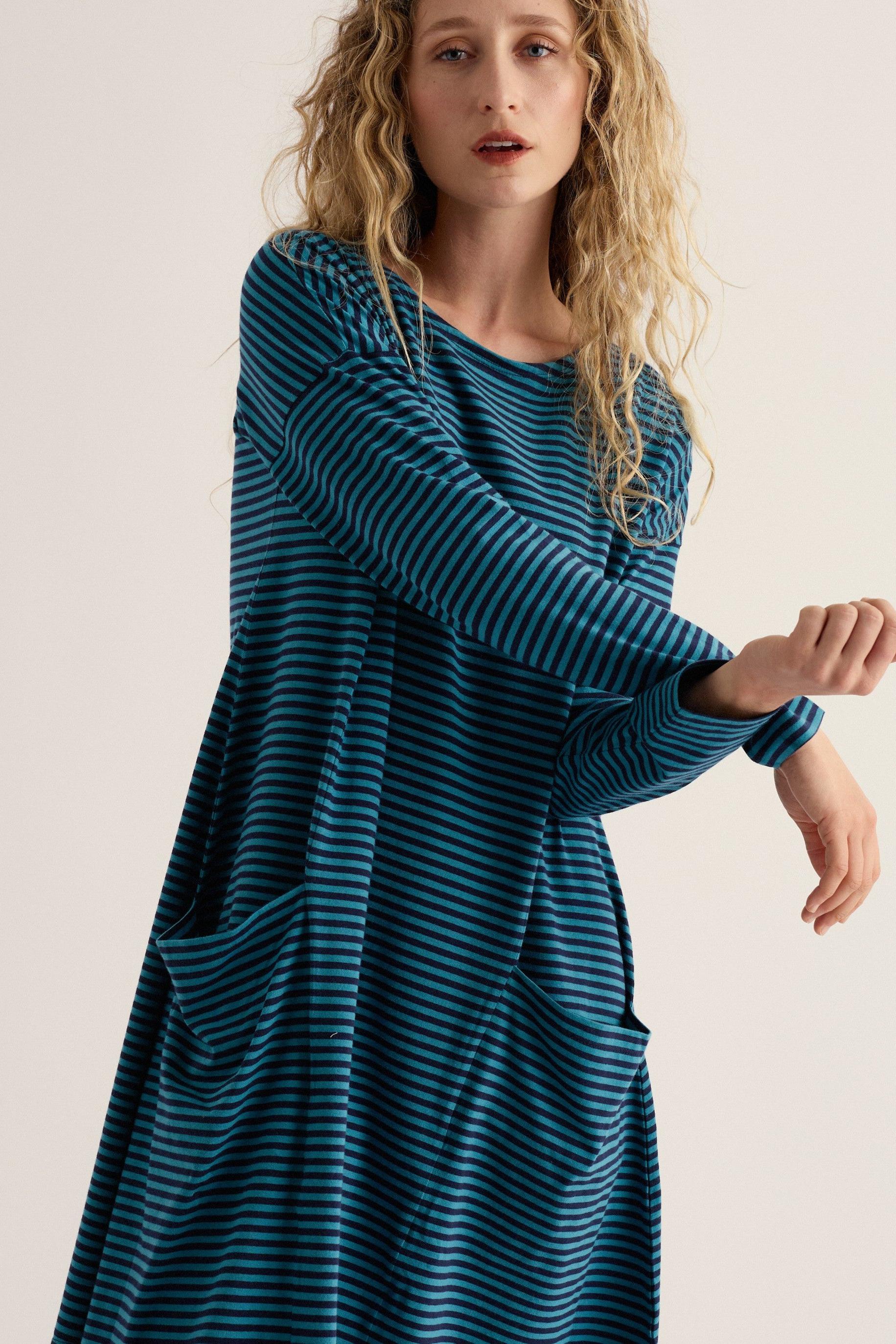 A woman wearing a long-sleeve, loose-fitting dress with horizontal teal and navy stripes, posed against a plain light beige studio background.
