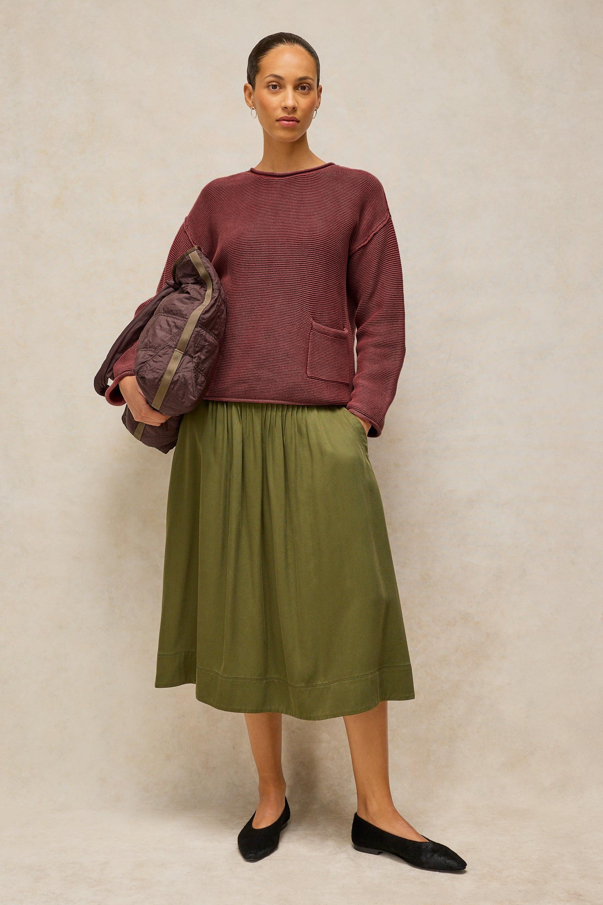 A woman stands against a textured beige studio backdrop wearing a dusty rose knit sweater, an olive green midi skirt, and black flats, while holding a dark patterned clutch.
