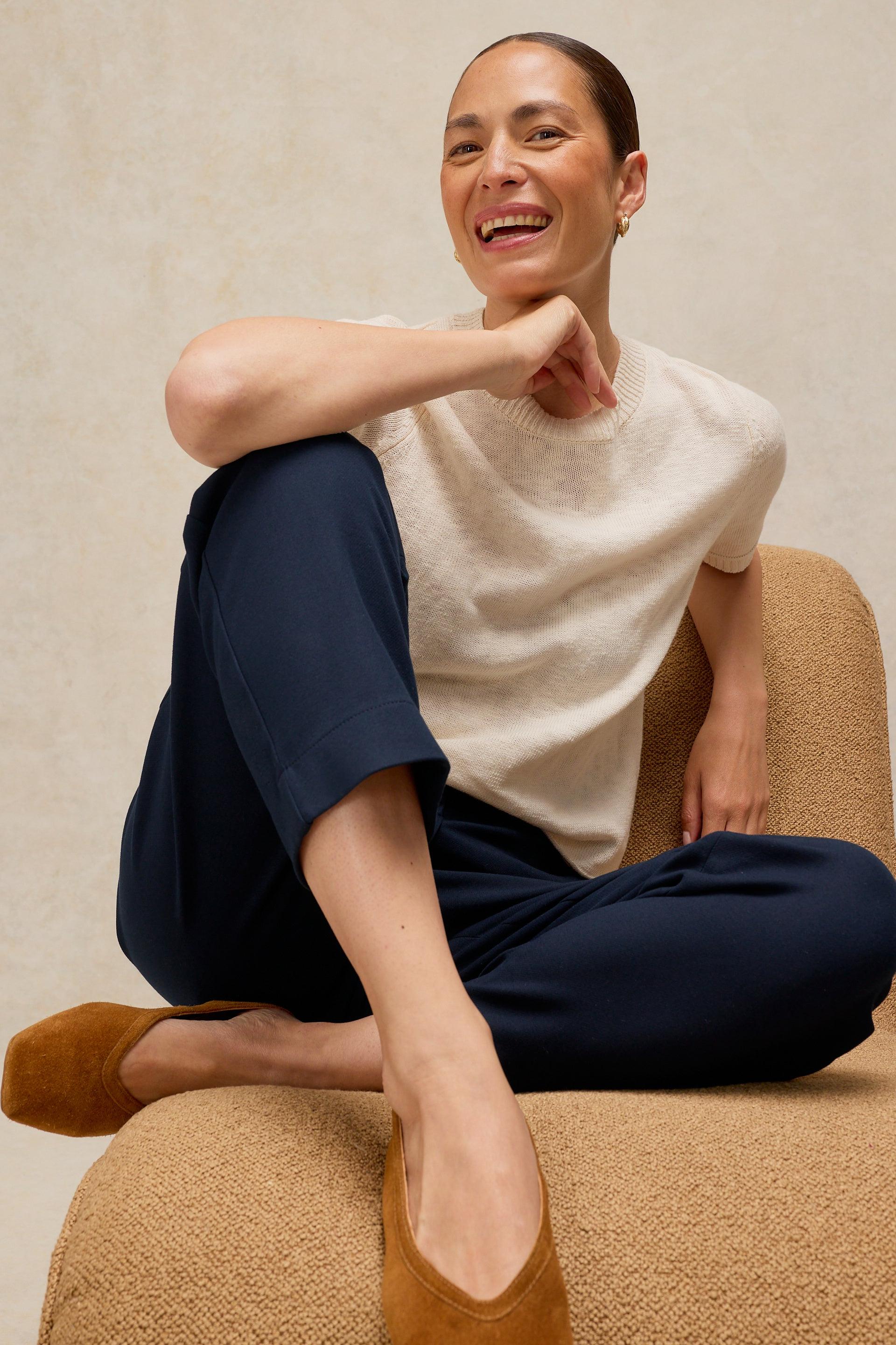 A woman sitting on a textured beige armchair, wearing a cream knit short-sleeve top, navy blue cropped trousers, and tan suede flats against a neutral beige background.