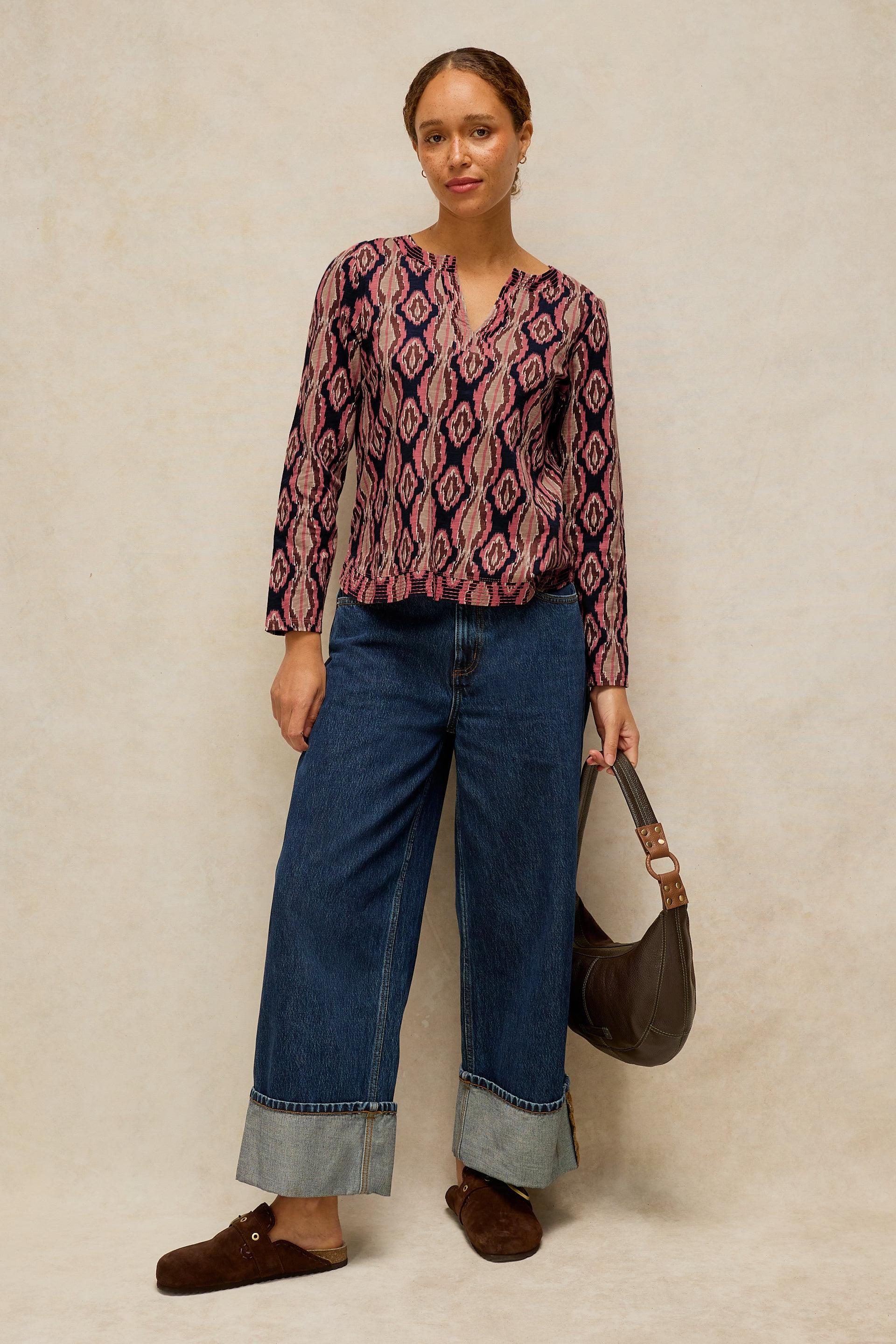 A woman stands against a textured beige studio backdrop wearing a long-sleeve V-neck blouse with a pink, purple, and dark geometric diamond pattern, paired with wide-leg blue jeans, brown slip-on shoes, and holding a dark brown shoulder bag.