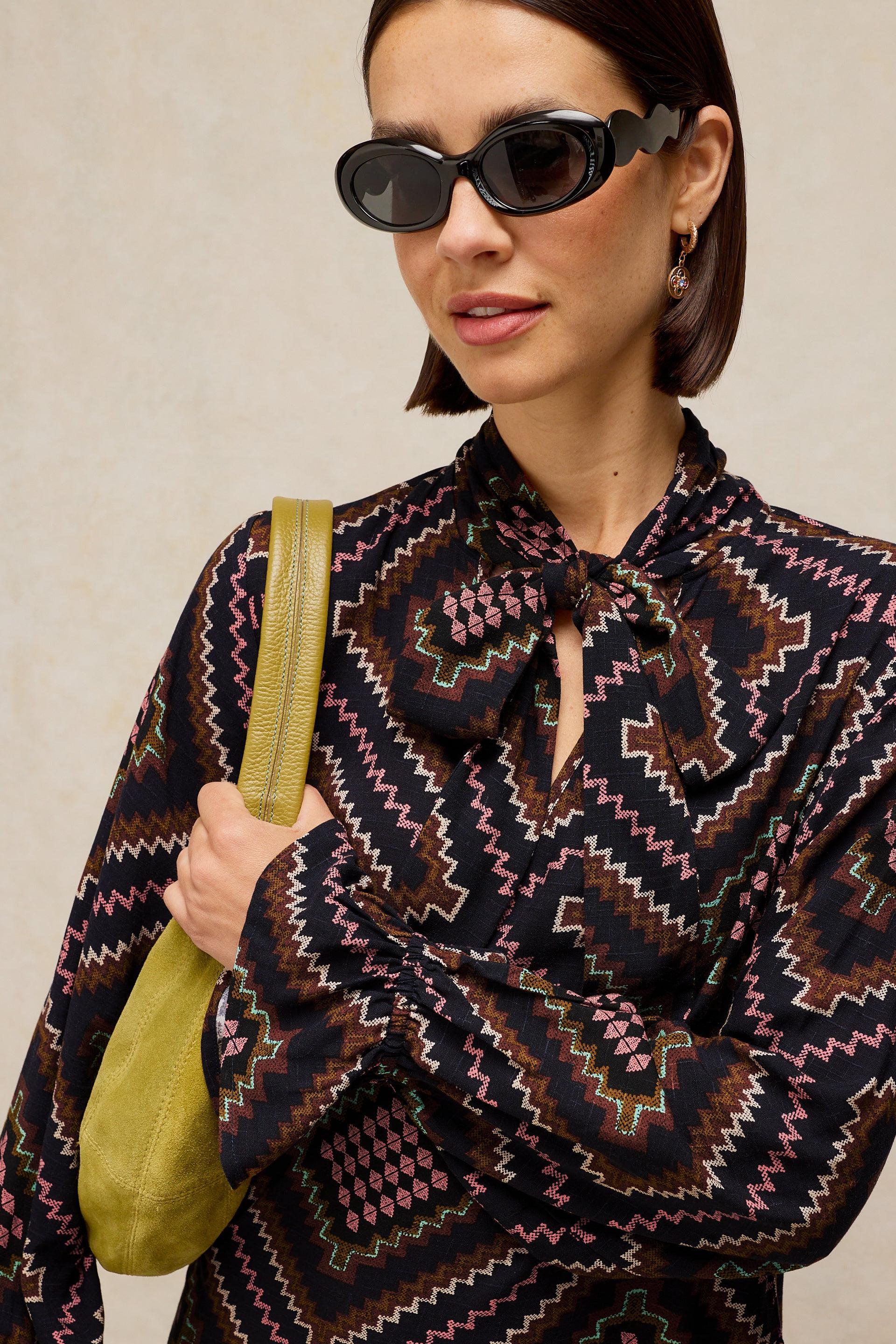 Woman wearing a patterned dress and sunglasses, holding a green bag against a neutral background