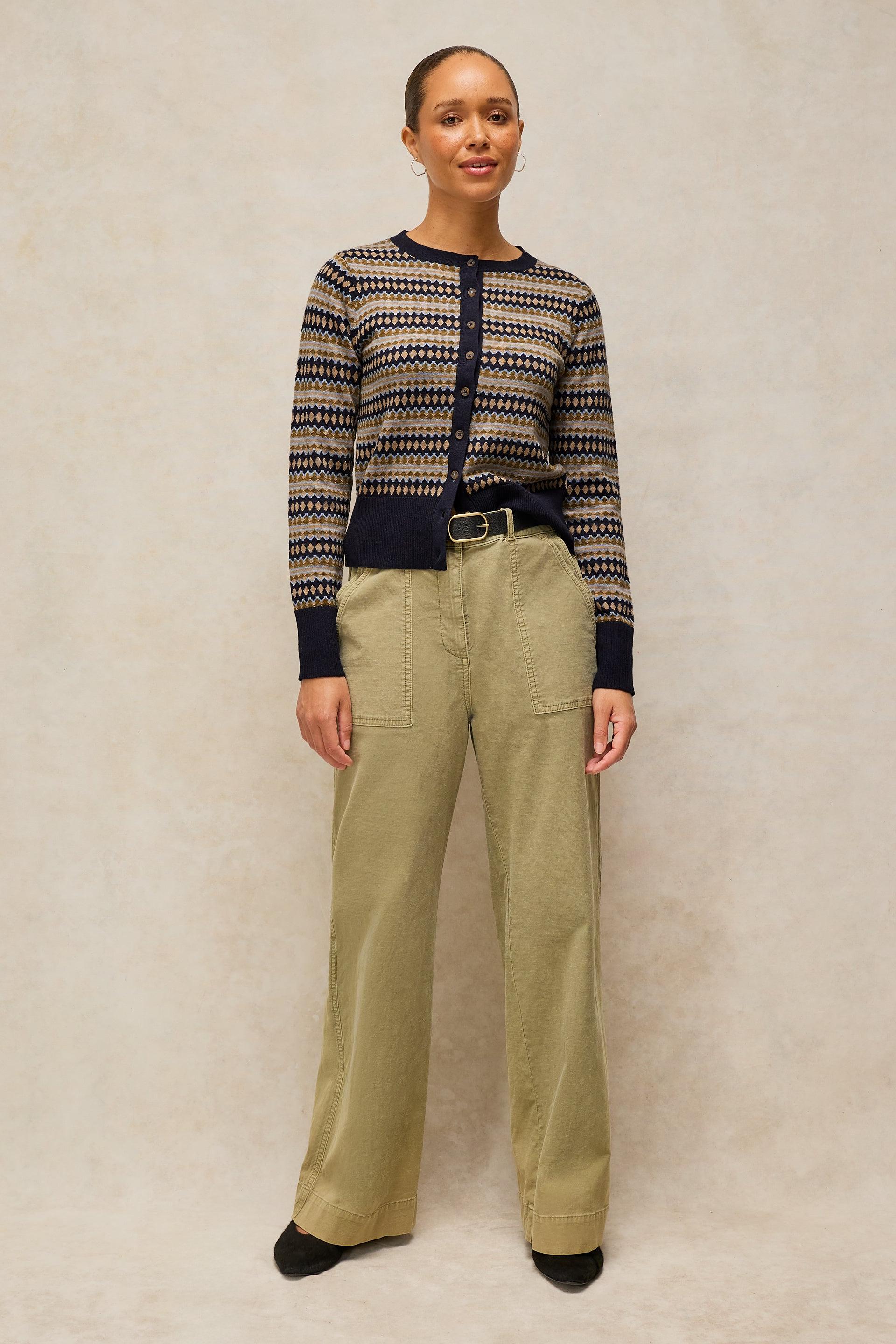 A woman stands against a textured beige studio background wearing a navy and beige patterned knit cardigan, olive green wide-leg trousers, and black shoes.