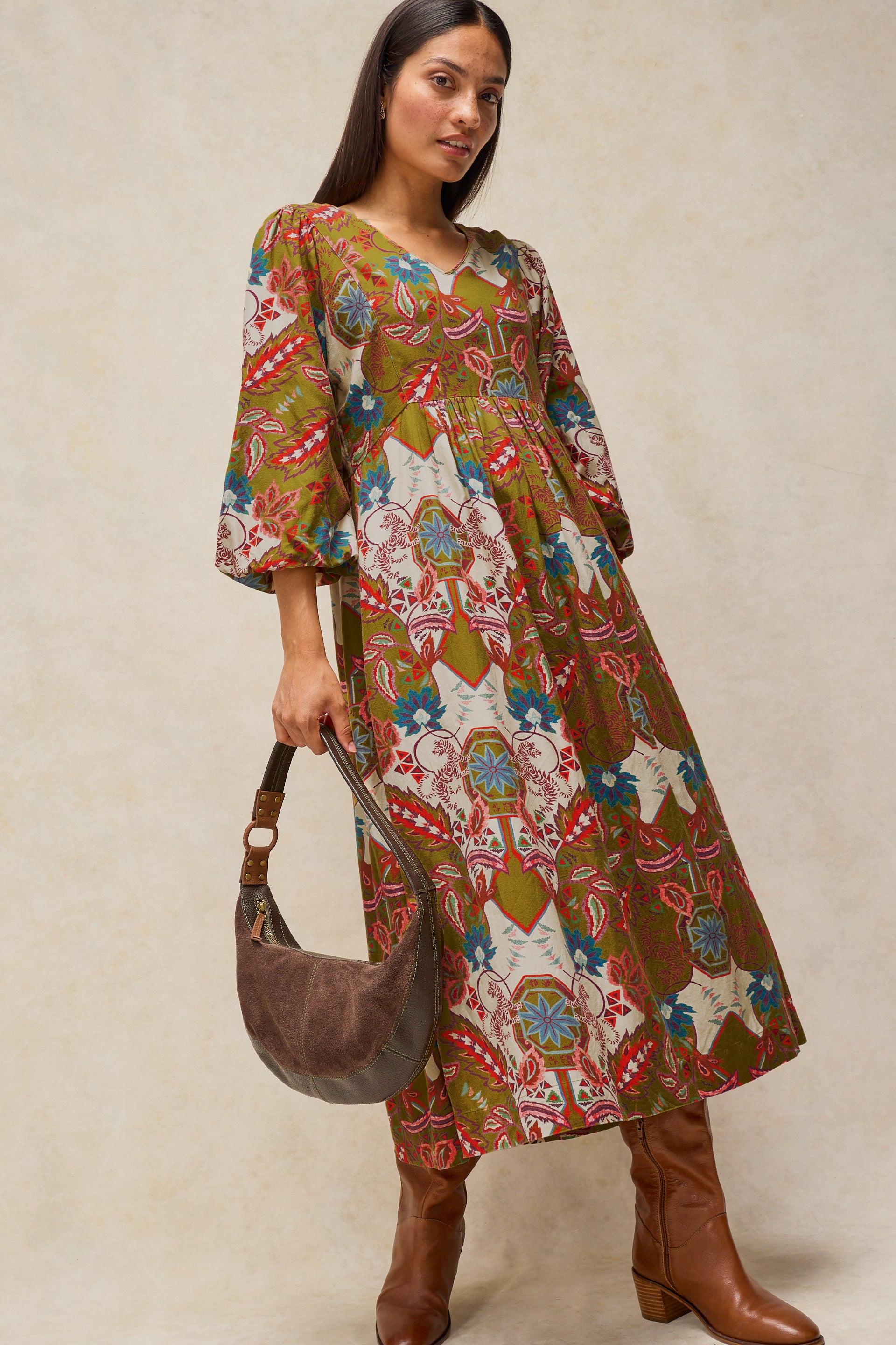 A woman models a bohemian-style midi dress with a multicolor abstract print in green, brown, red, and cream. She is holding a brown suede crescent shoulder bag and wearing brown leather boots, set against a neutral beige studio background.