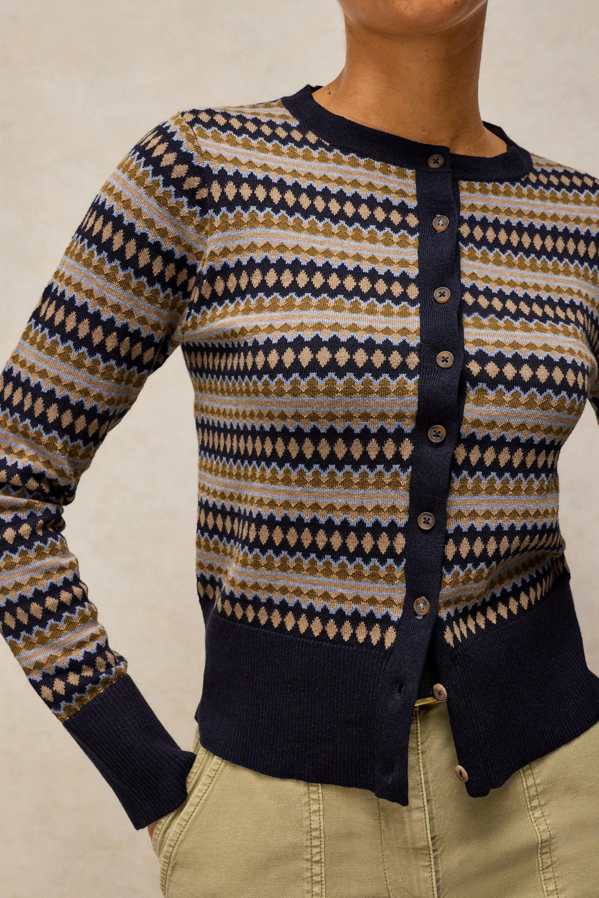 A close-up of a woman wearing a long-sleeve button-front cardigan with a navy, beige, and brown geometric diamond pattern, paired with khaki pants.